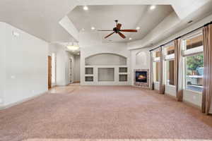 Spacious living area featuring a tray ceiling with recessed lighting and a ceiling fan, a built-in media center with arched alcoves, a gas fireplace with stone surround, and multiple large windows with window treatments