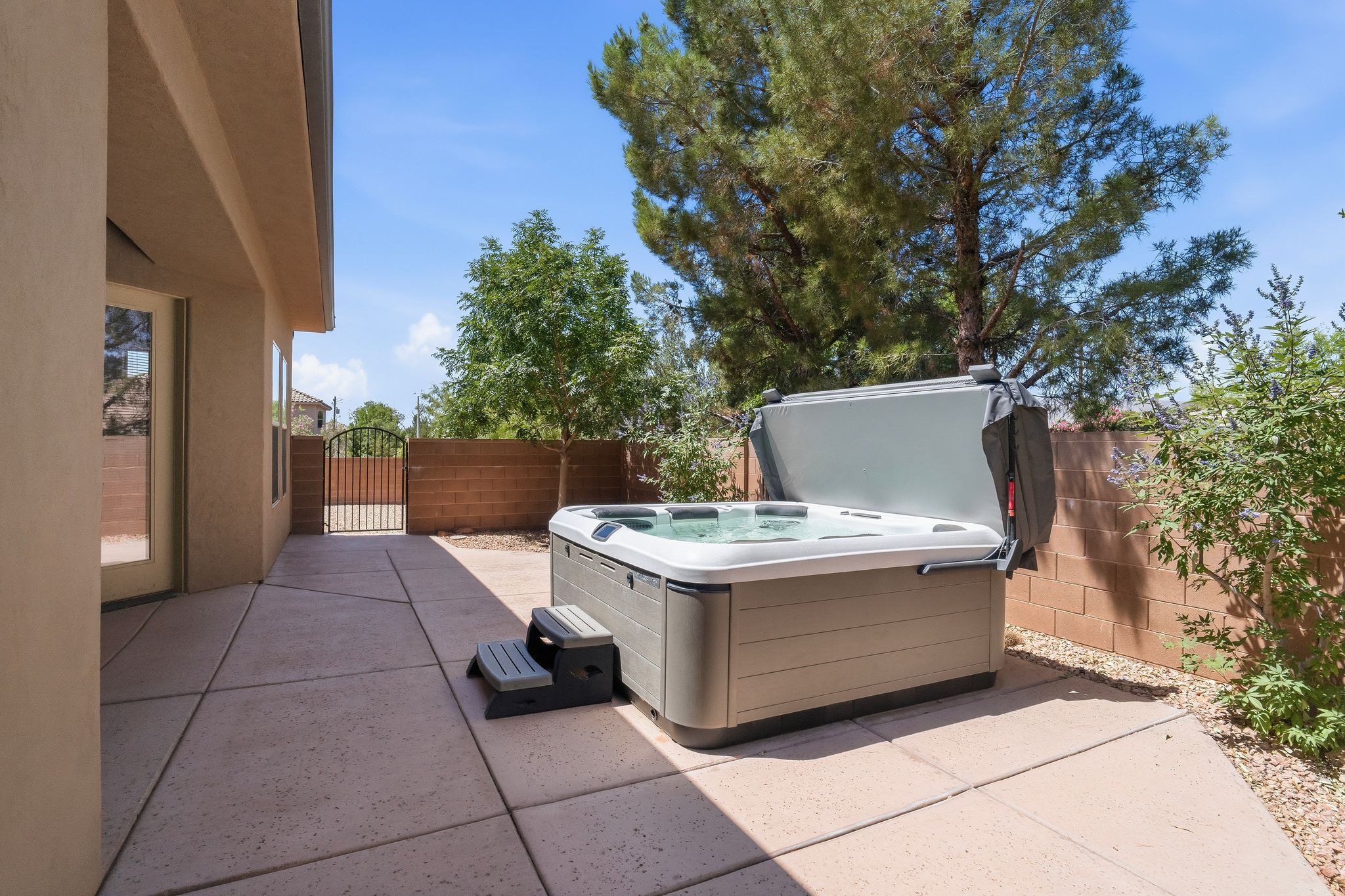 Outdoor patio featuring a hot tub, concrete paving, a block wall perimeter, mature trees, and a wrought iron gate