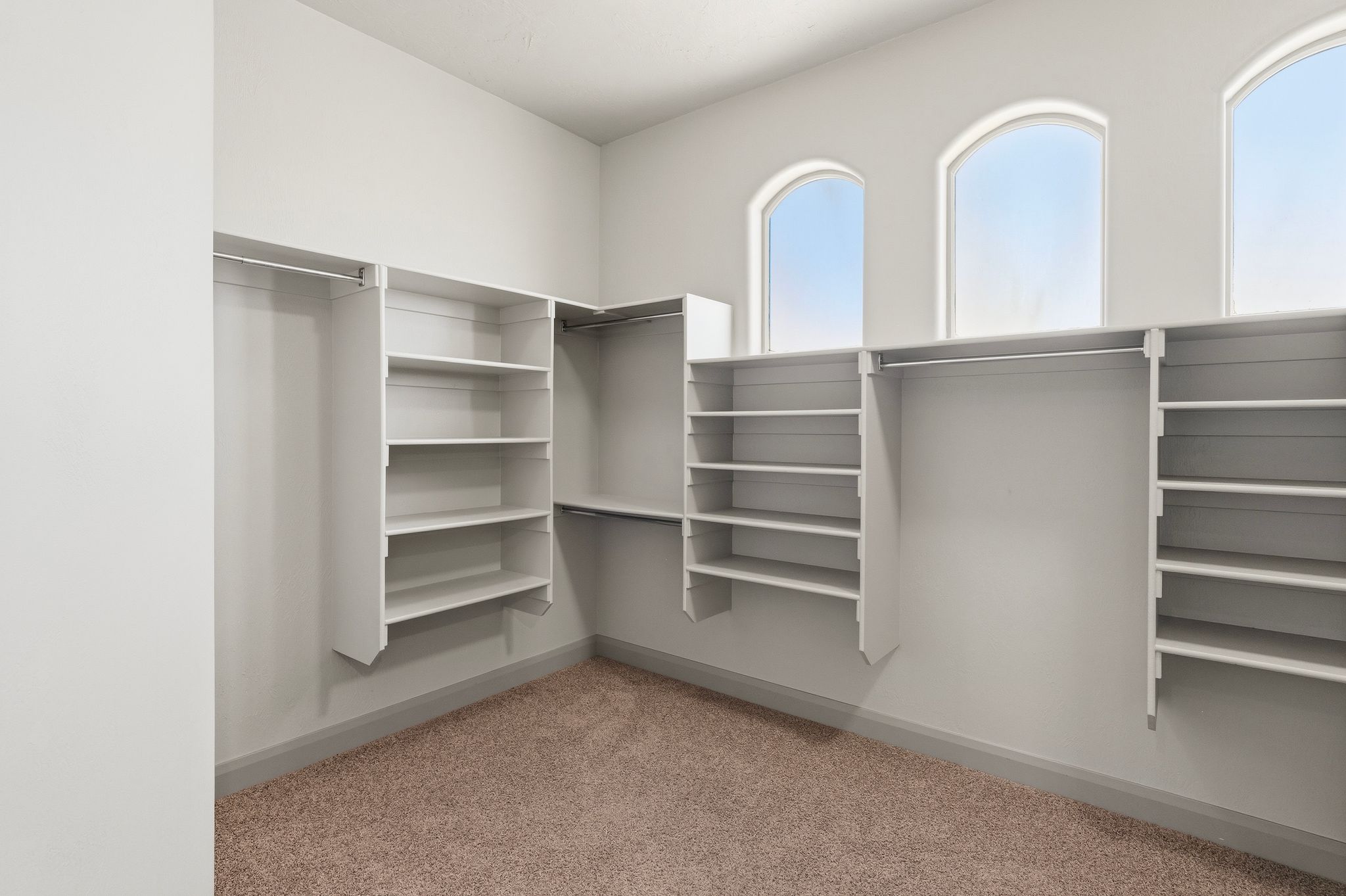 Primary suite walk-in closet featuring built-in shelving, multiple hanging rods, three arched windows, and light brown carpeting