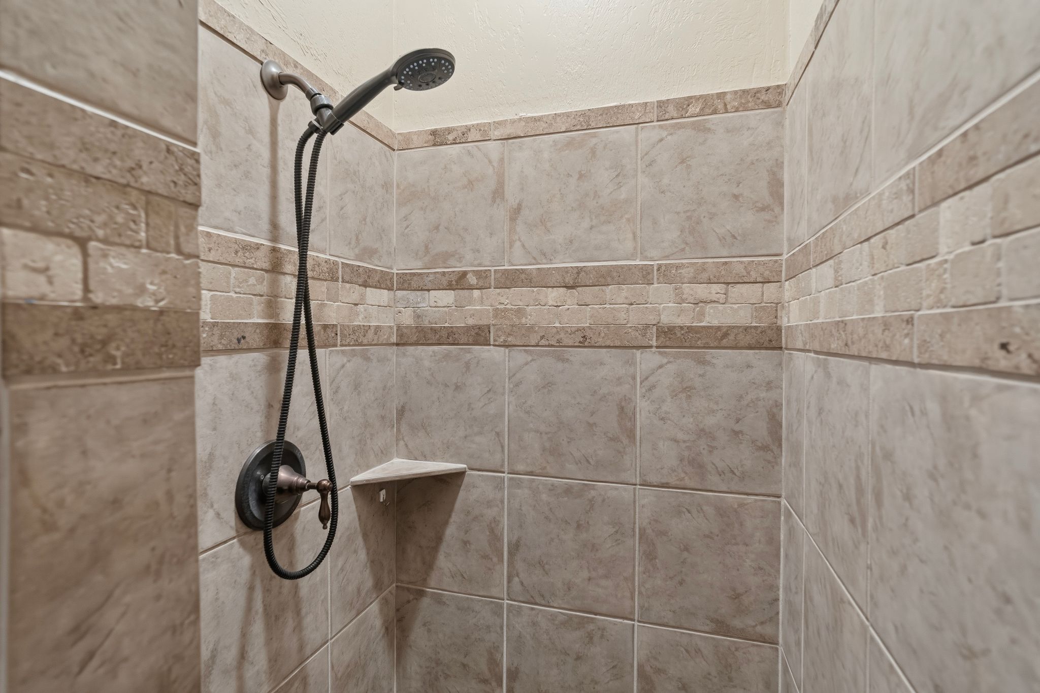 Shower stall featuring a handheld showerhead with bronze finish fixtures, a corner shelf, and neutral-toned ceramic tile with a decorative tile band