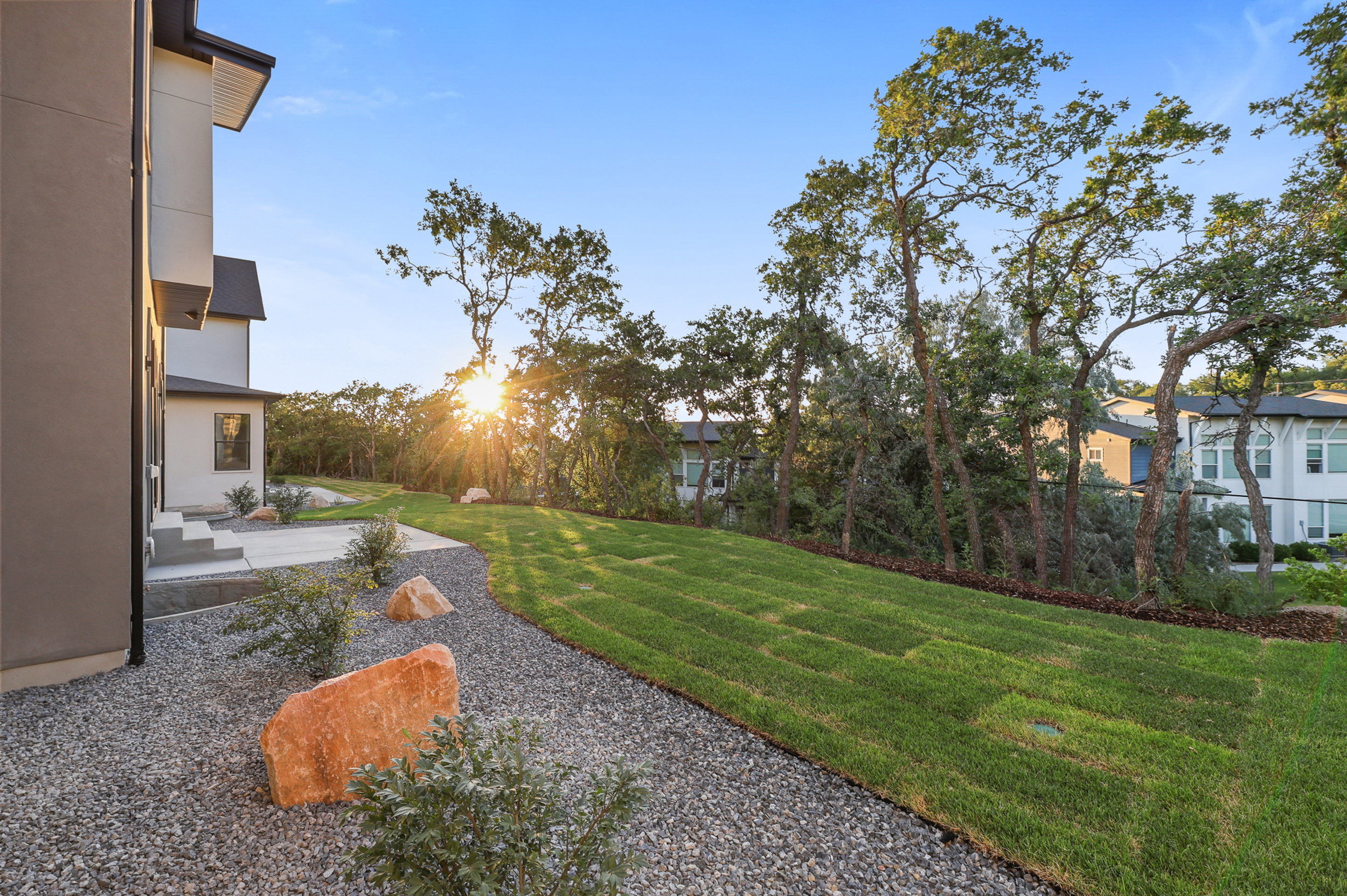 Expansive green space featuring a sloped lawn, mature trees, and a gravel-covered area with decorative boulders