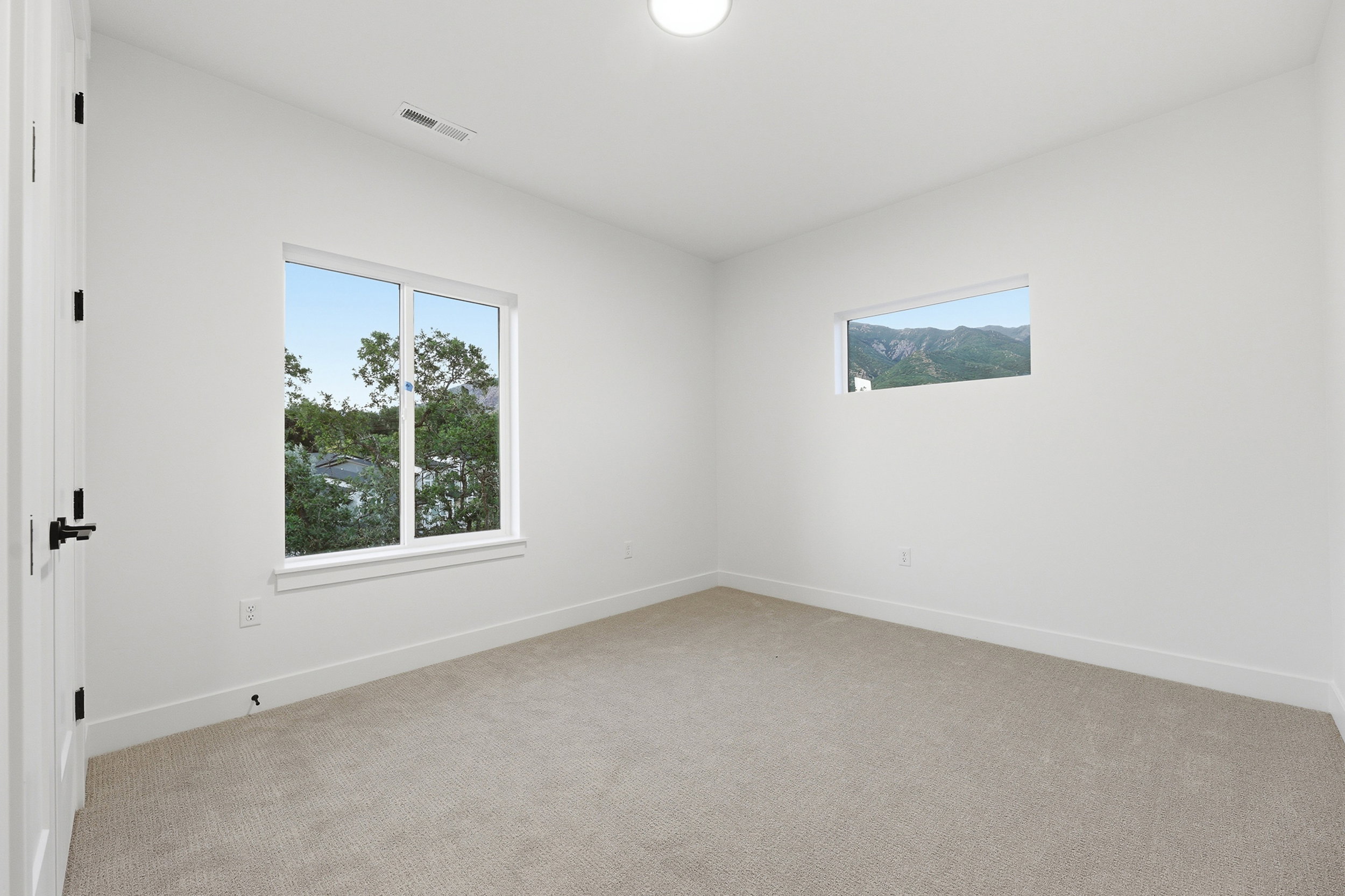 Spacious room featuring neutral carpeting, white walls, two windows with outdoor scenery, and a modern ceiling light fixture