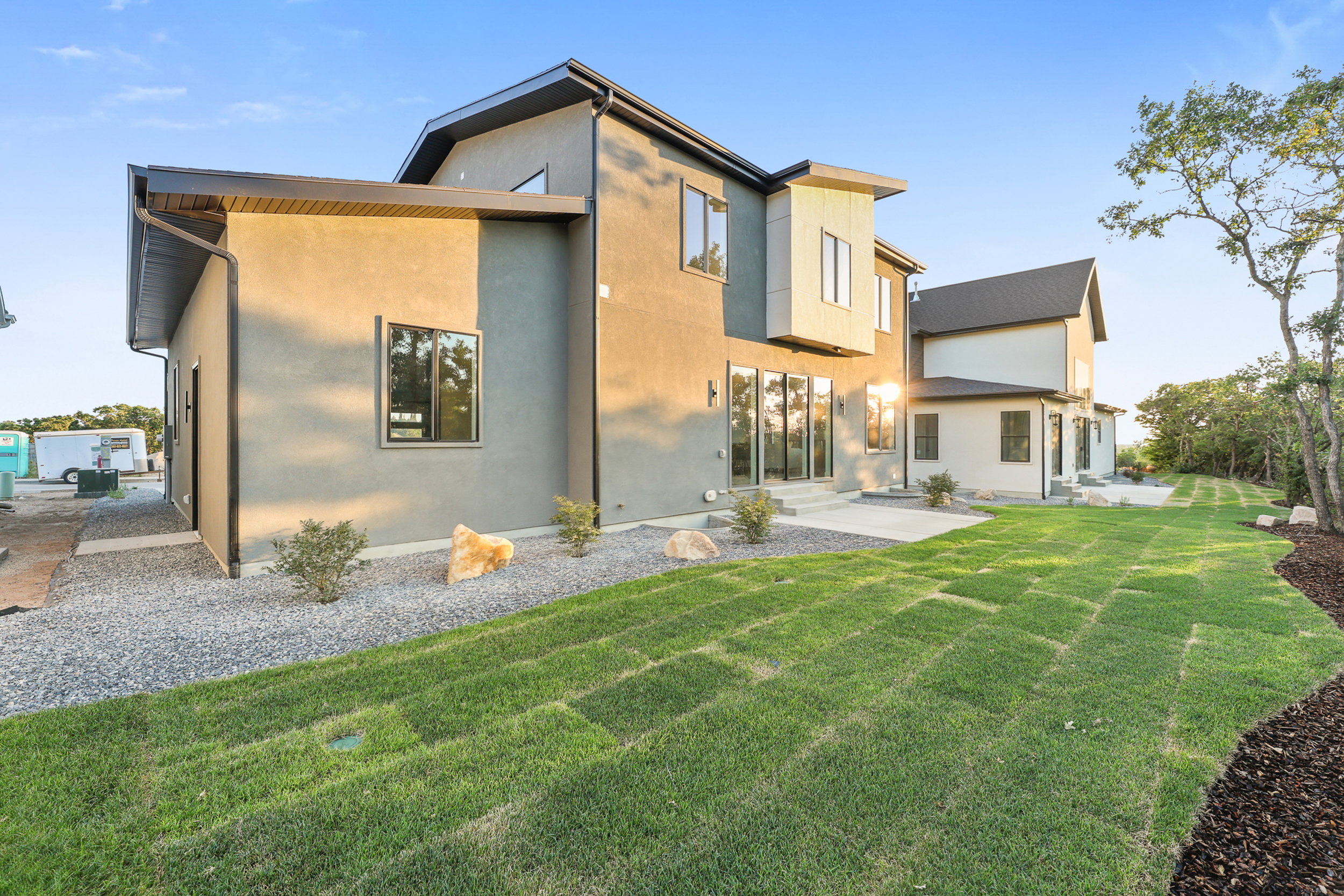 Contemporary exterior featuring a stucco finish, multiple large windows, a concrete patio, and a sloped lawn with decorative rock landscaping