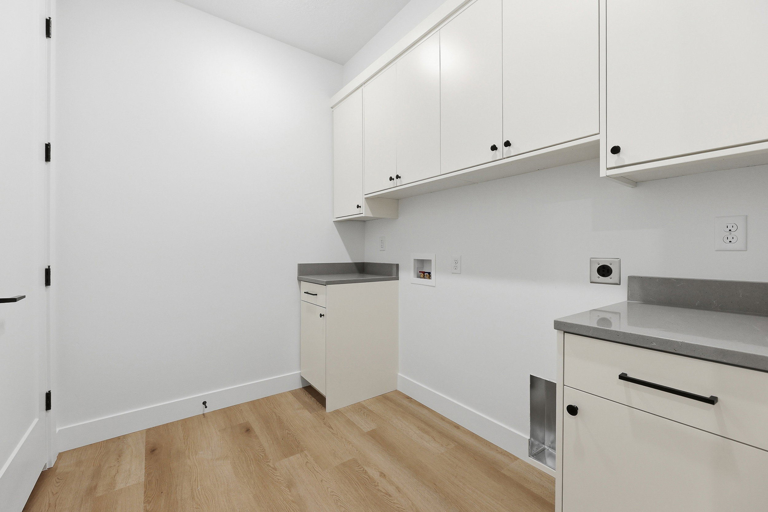 Utility room featuring wood-finish flooring, white cabinetry with black hardware, light gray countertops, and a white door with black hardware