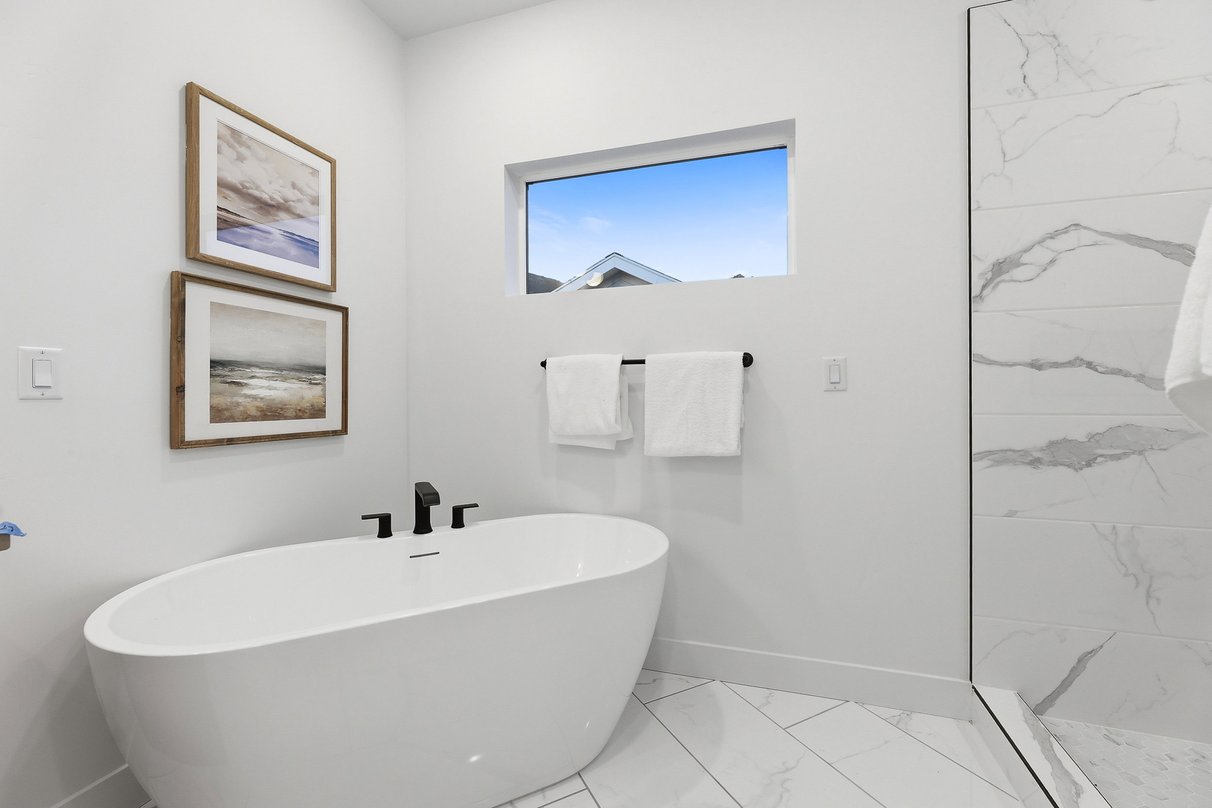 Freestanding soaking tub with matte black fixtures, set against a backdrop of white walls and large format marble-look tile flooring