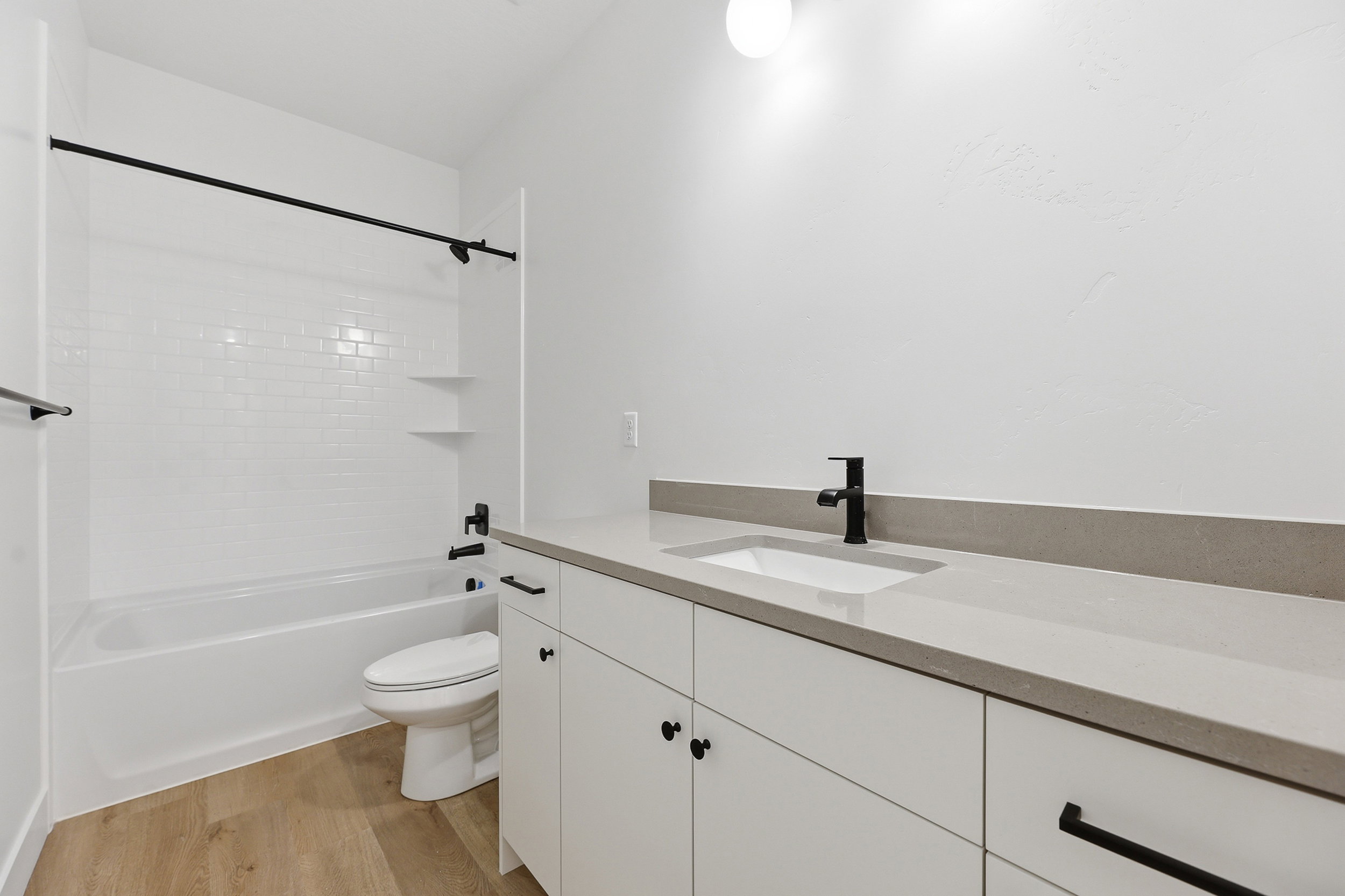 Modern bathroom featuring a white subway tile tub surround, matte black fixtures, a contemporary vanity with a light-toned countertop, an undermount sink, and wood-finish flooring