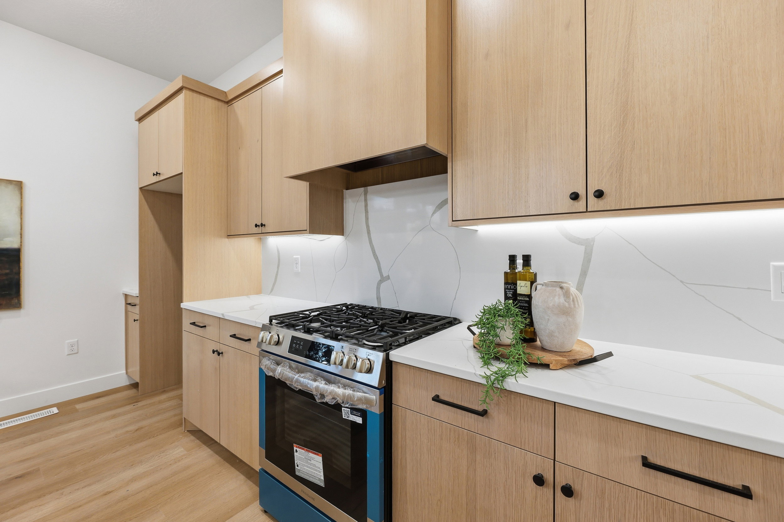 Kitchen featuring a stainless steel gas range, light wood-finish cabinetry, white countertops, a veined white backsplash, and under-cabinet lighting