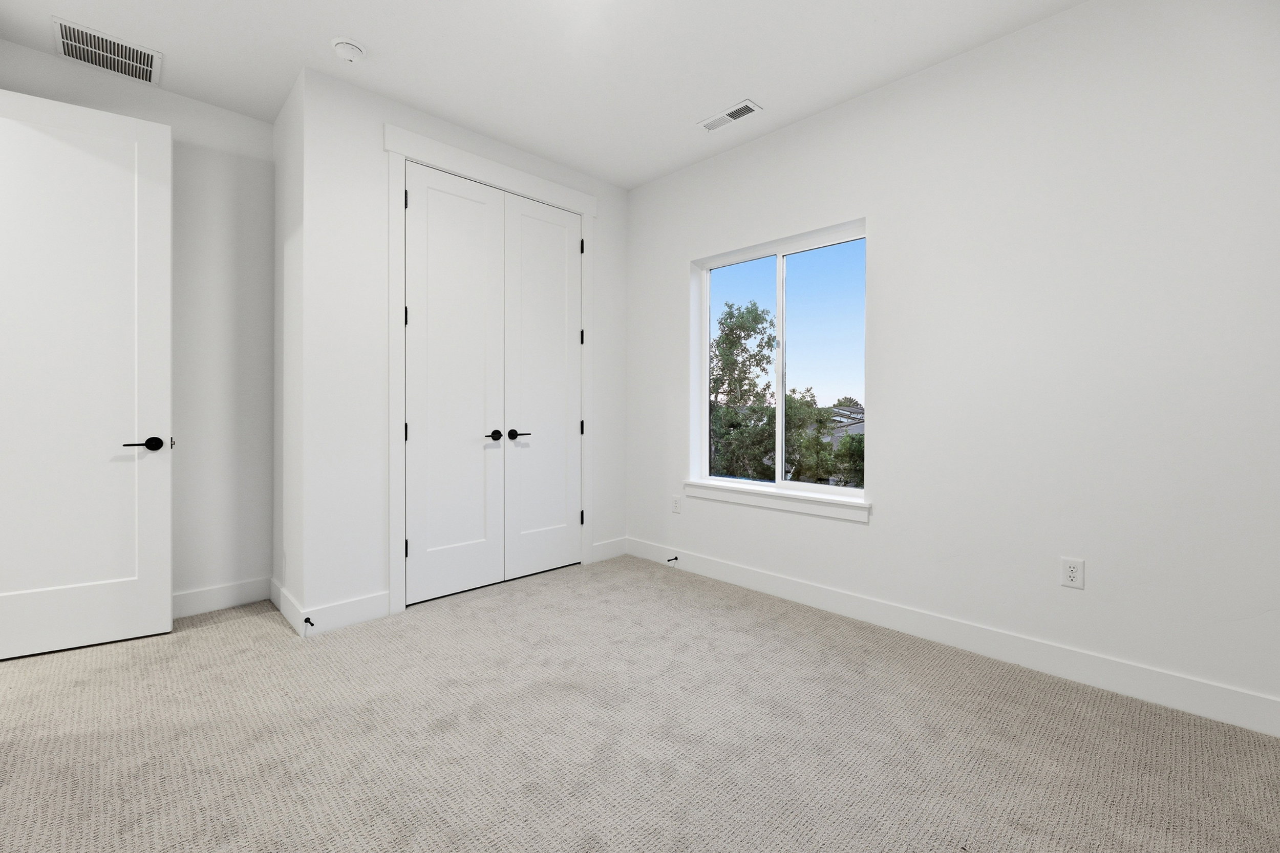 Bright room featuring light gray carpet, white walls, a single window with white trim, and a double-door closet with black hardware