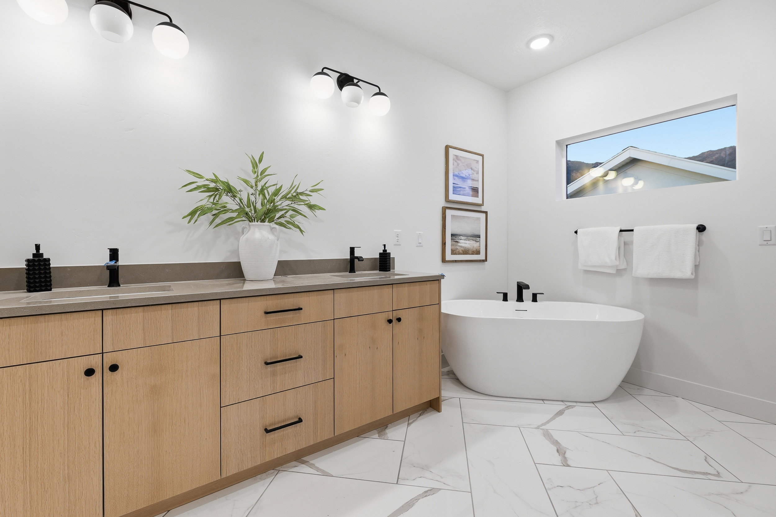 Contemporary bathroom featuring a freestanding soaking tub, wood-finish double vanity with stone countertops, and a rectangular transom window