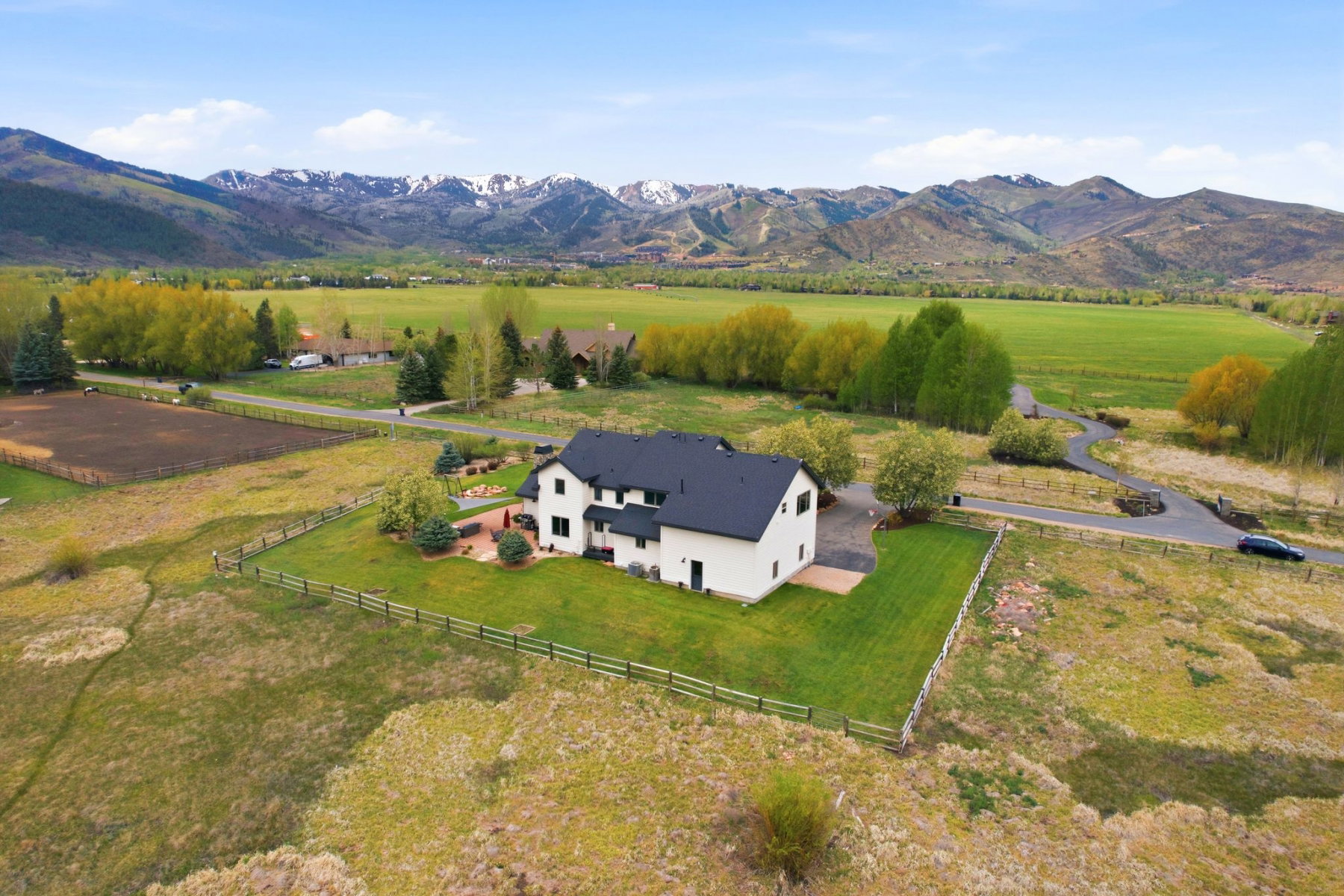 Contemporary residence featuring a black roof and white siding, a manicured lawn, an outdoor patio space, and a paved driveway, all set against a backdrop of distant mountains and open fields