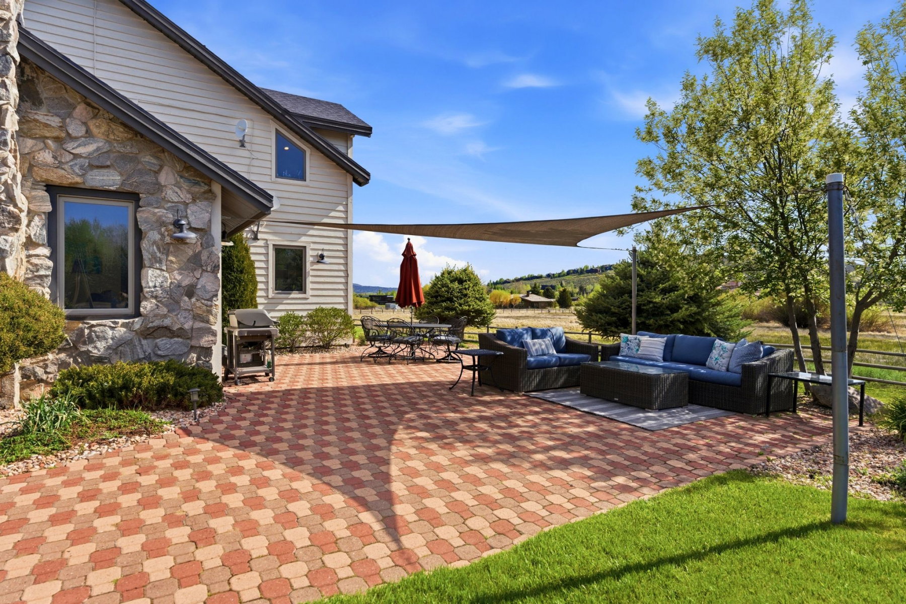 Expansive paver patio with a shade sail, featuring stone and siding exterior elements, surrounded by mature landscaping and open grounds