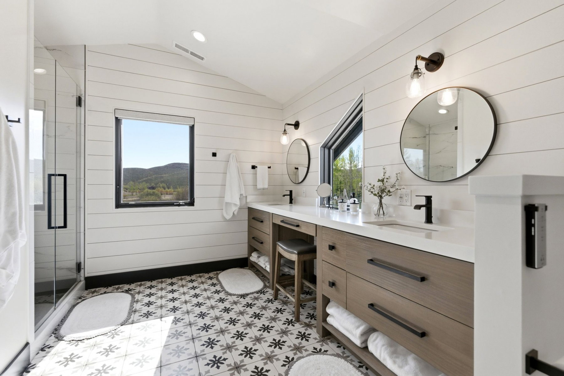 Spacious bathroom featuring a dual vanity with light wood-finish cabinetry and white countertops, a glass-enclosed shower, patterned floor tile, white shiplap walls, and two windows