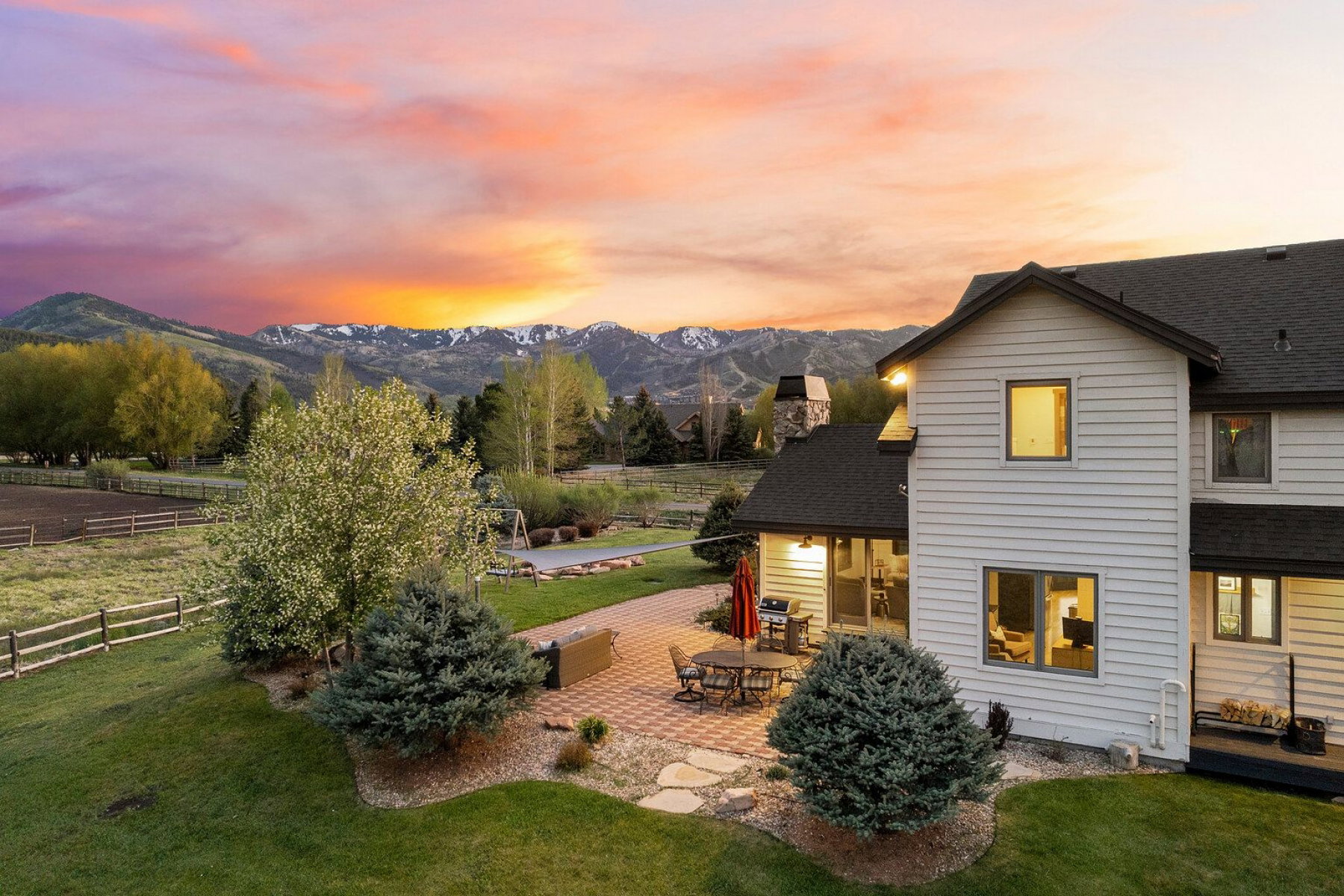 Rear exterior featuring an expansive paved patio, lush green lawn, mature trees, and a mountain backdrop