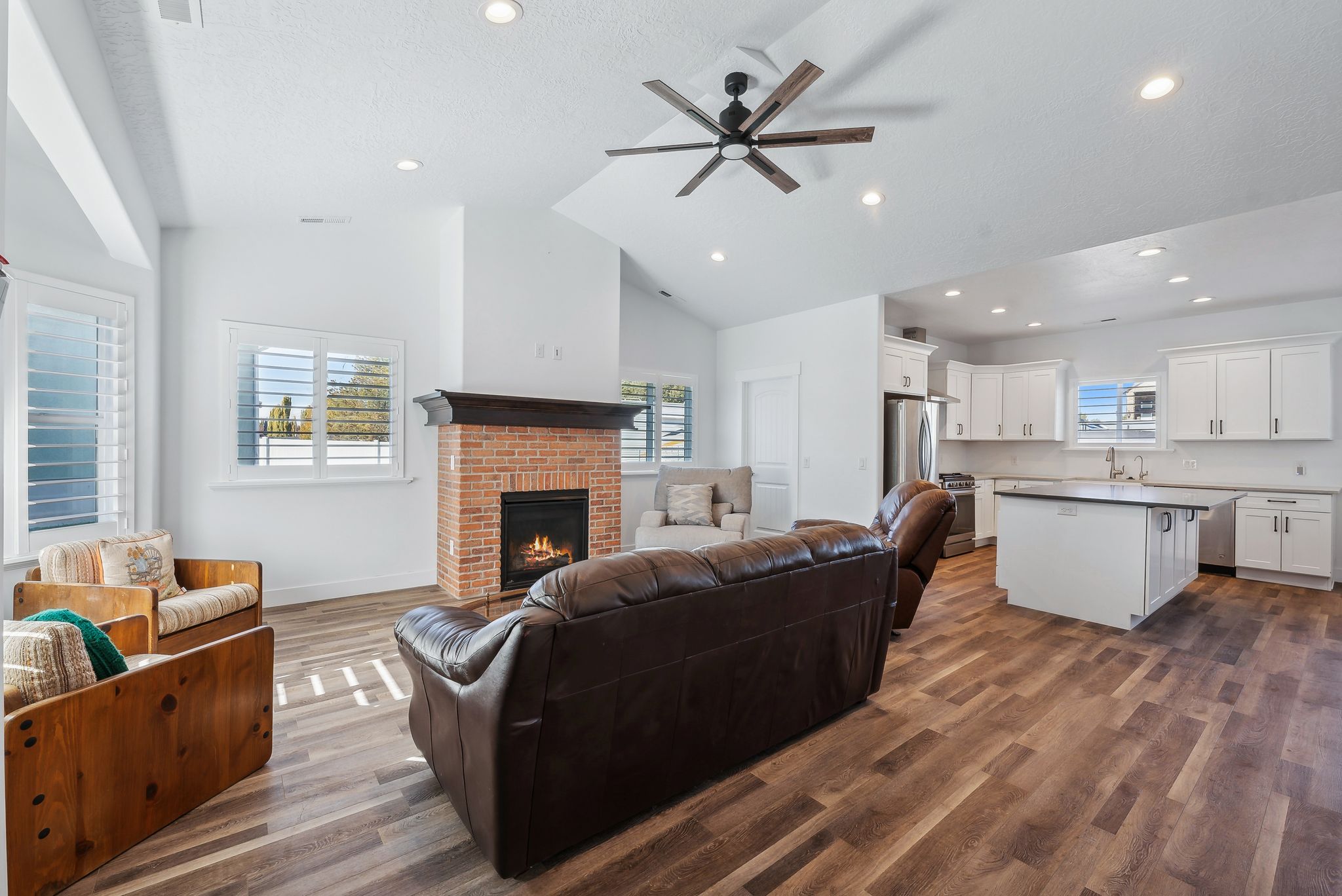 Custom built ADU/Open-concept living space featuring wood-finish flooring, a brick-surround fireplace with a dark wood mantle, and white plantation shutters on multiple windows