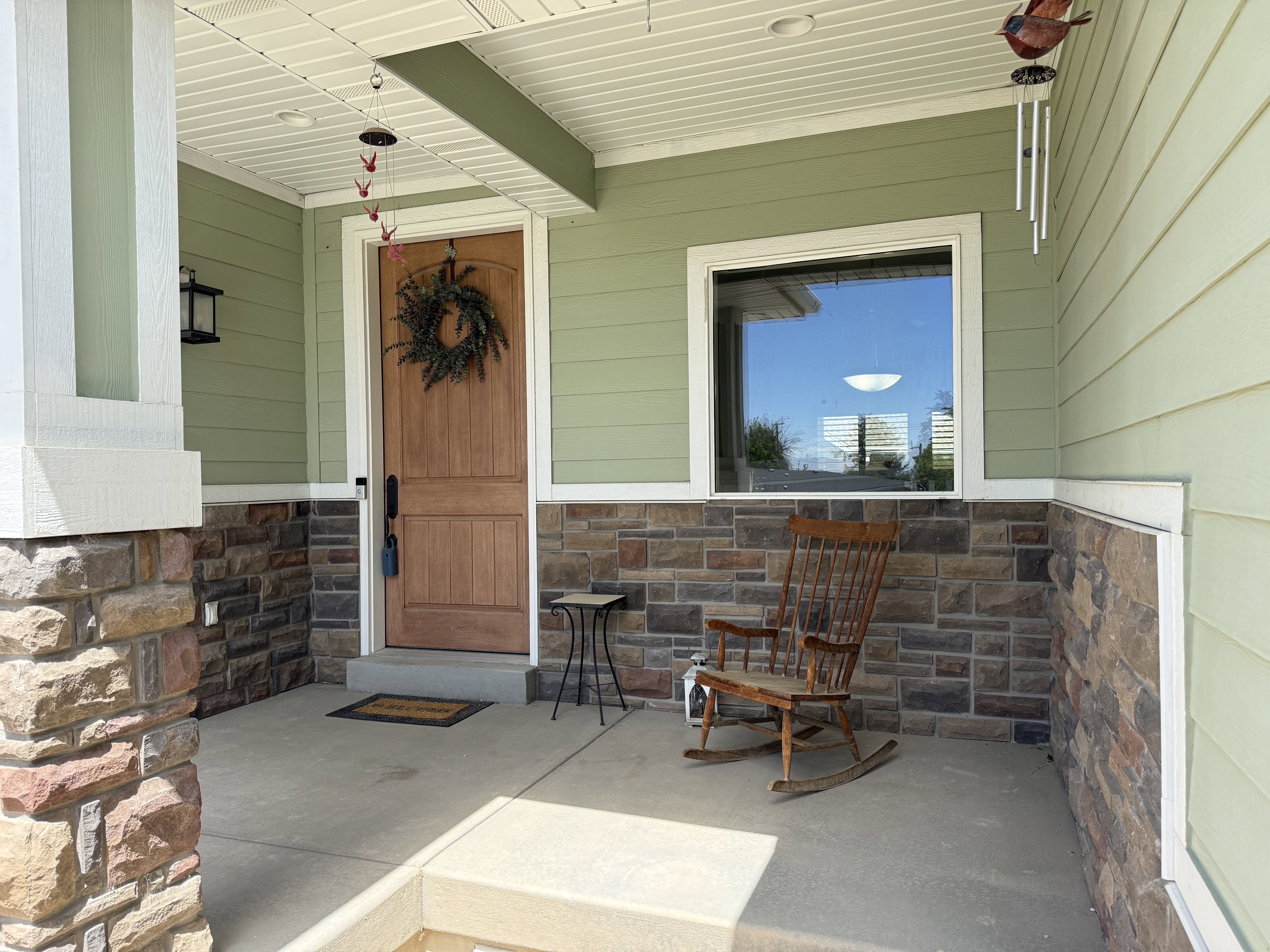Inviting covered porch featuring a natural wood-finish entry door, stone veneer detailing, horizontal siding, exterior lighting fixture, and a large window