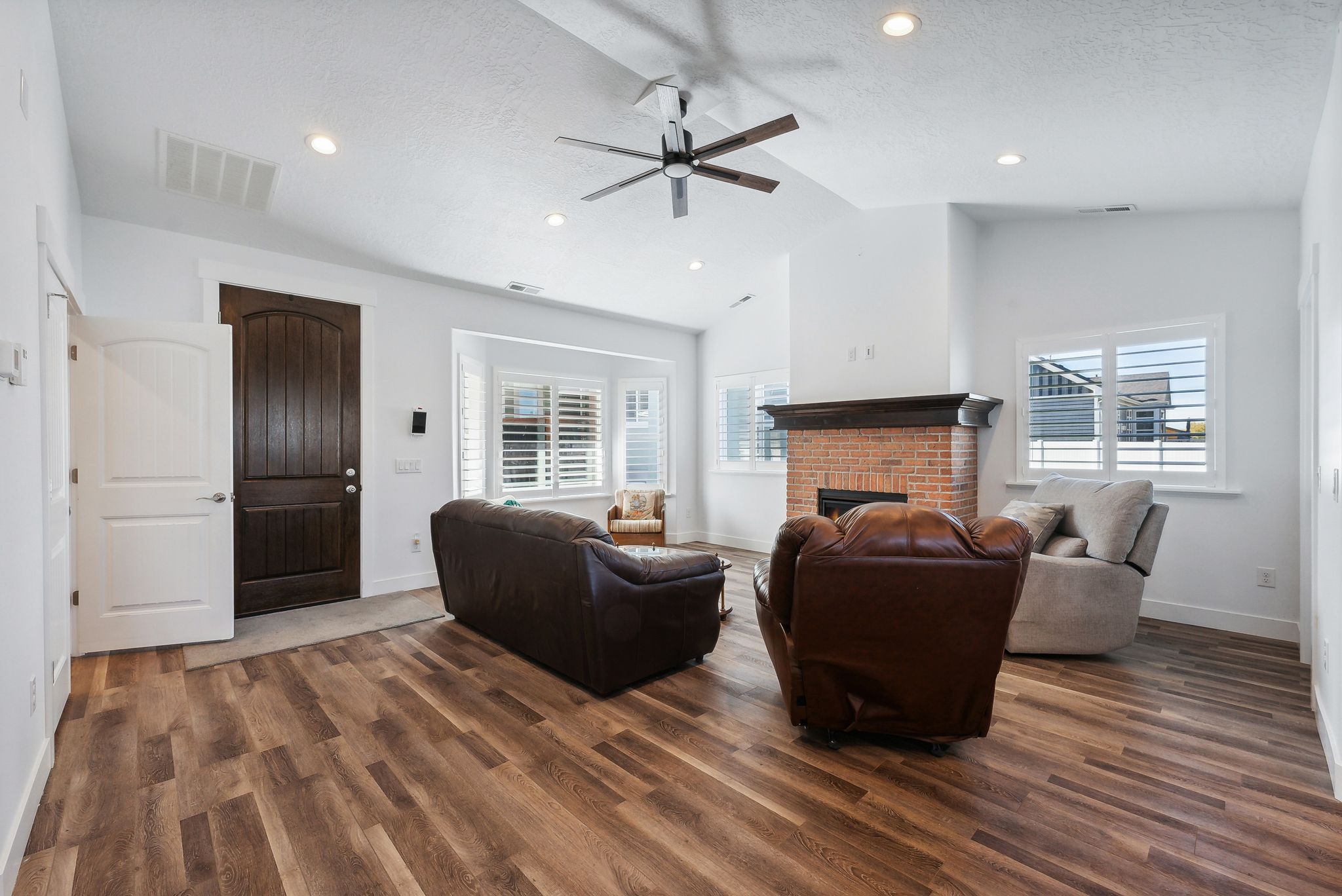 Custom built ADU/Spacious living area featuring wood-finish flooring, a brick fireplace with a dark wood mantel, and multiple windows with plantation shutters