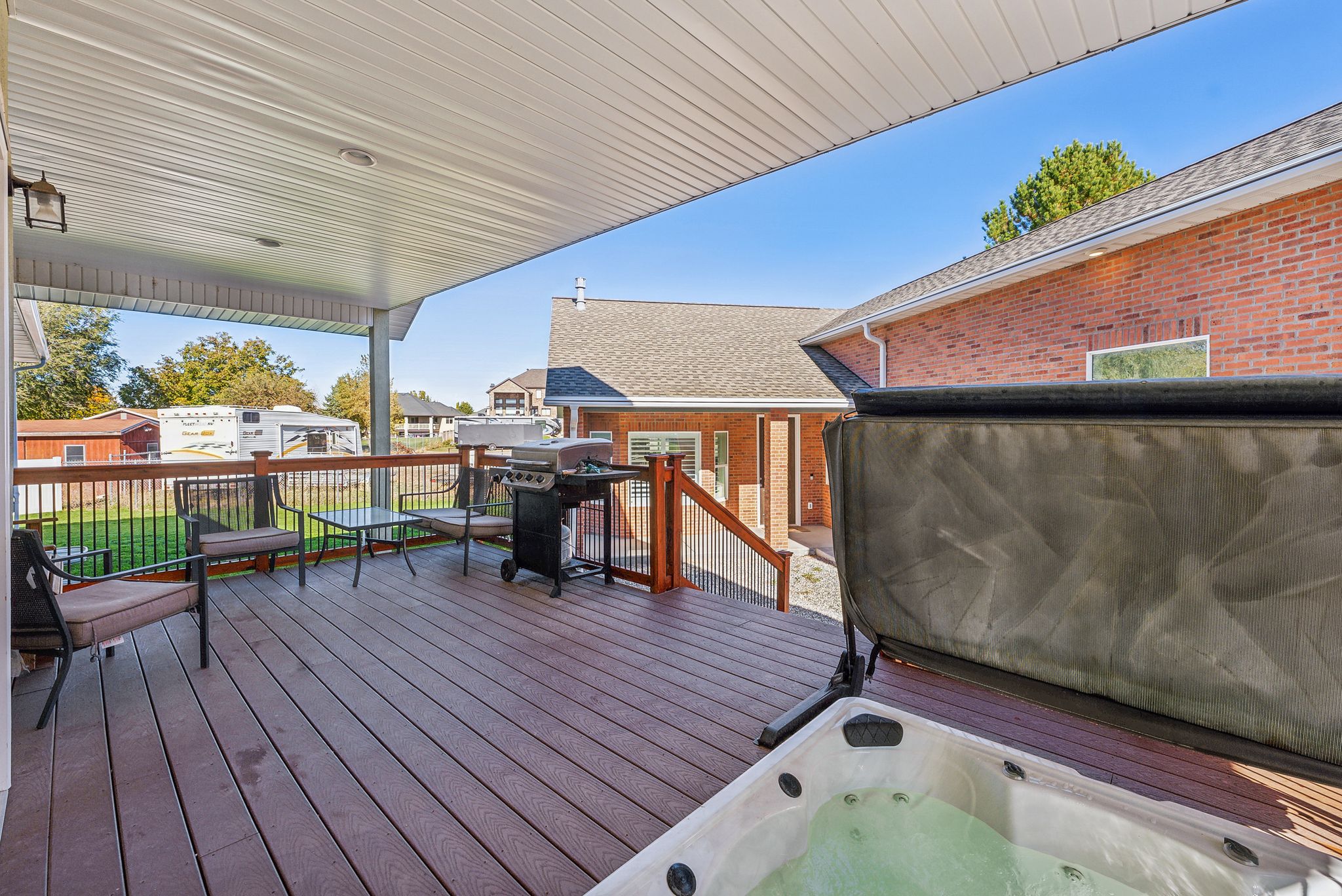 Spacious covered deck featuring composite decking, wood railing with black metal spindles, a built-in hot tub, and exterior lighting fixtures