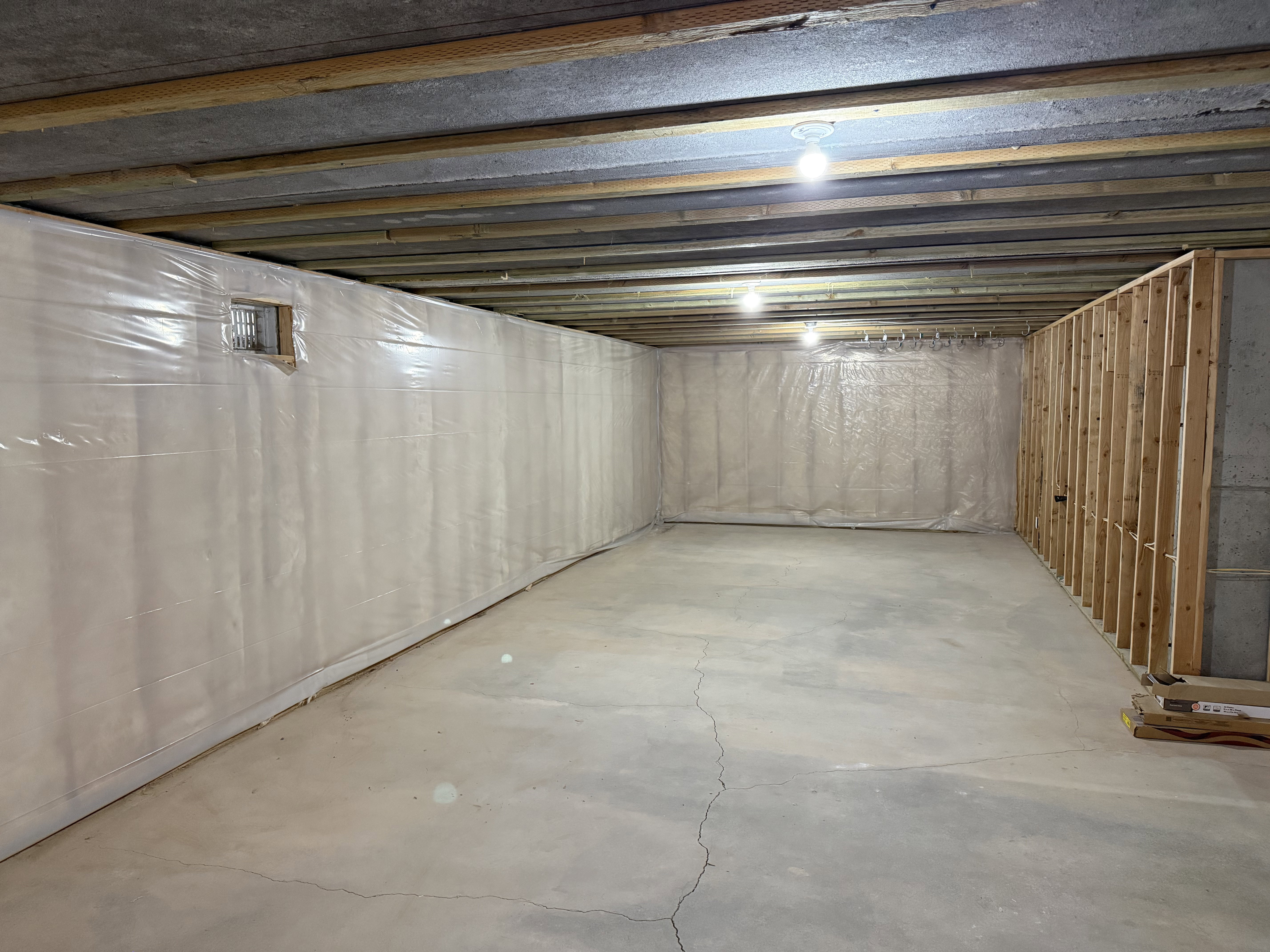 Unfinished basement featuring exposed ceiling joists, concrete flooring, vapor barrier wall covering, and partial wood framing for future development