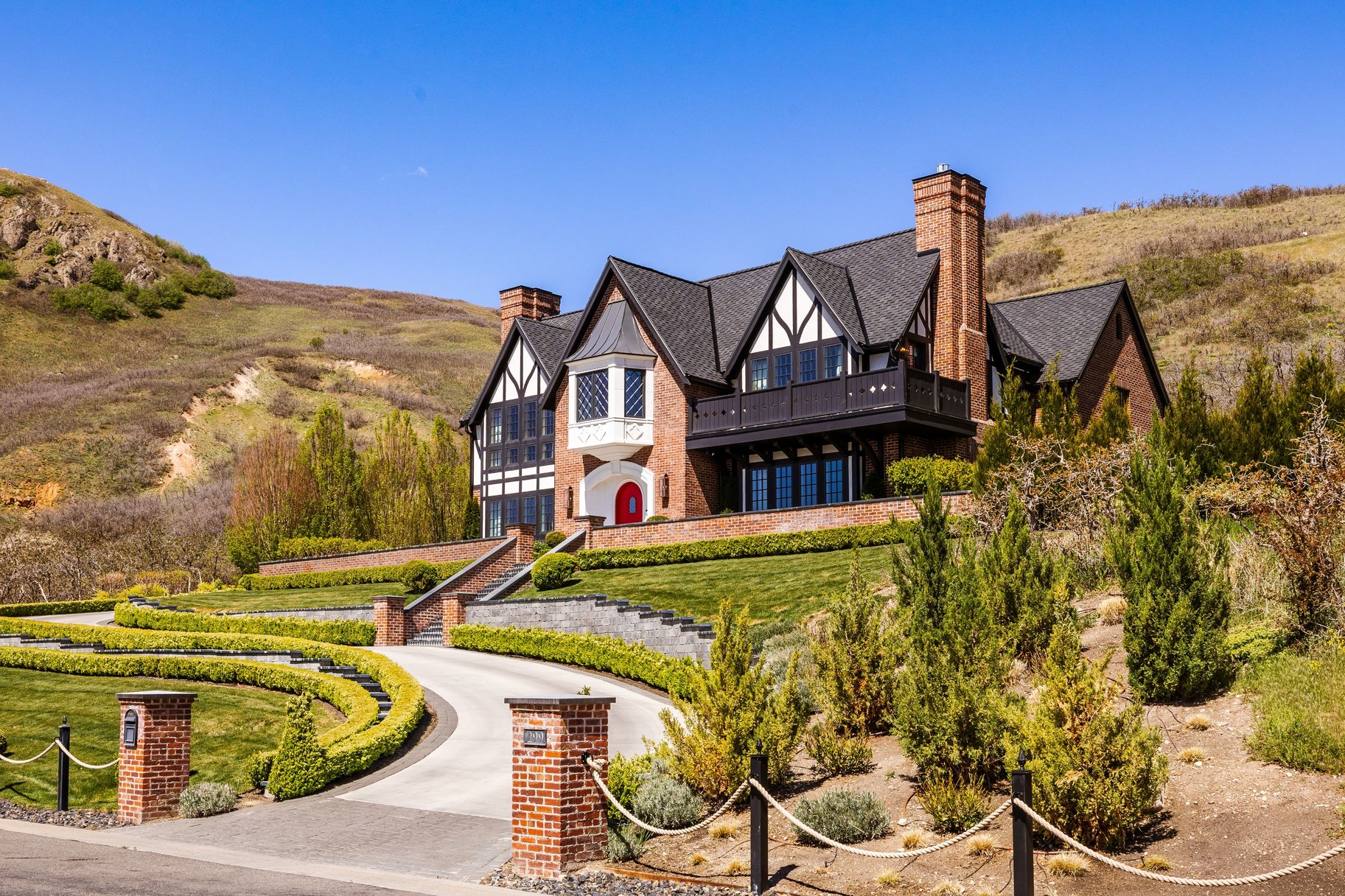 Brick and Tudor-style residence featuring a prominent red entry door, a multi-tiered front lawn, a winding paved driveway, and mature landscaping