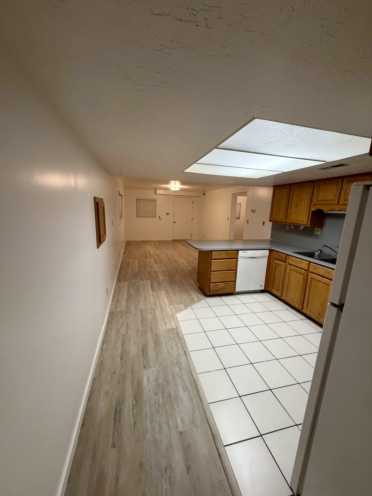 Open-concept kitchen and living area featuring wood-finish flooring in the main space, white ceramic tile in the kitchen, natural wood cabinetry, a built-in dishwasher, and a large overhead lighting panel