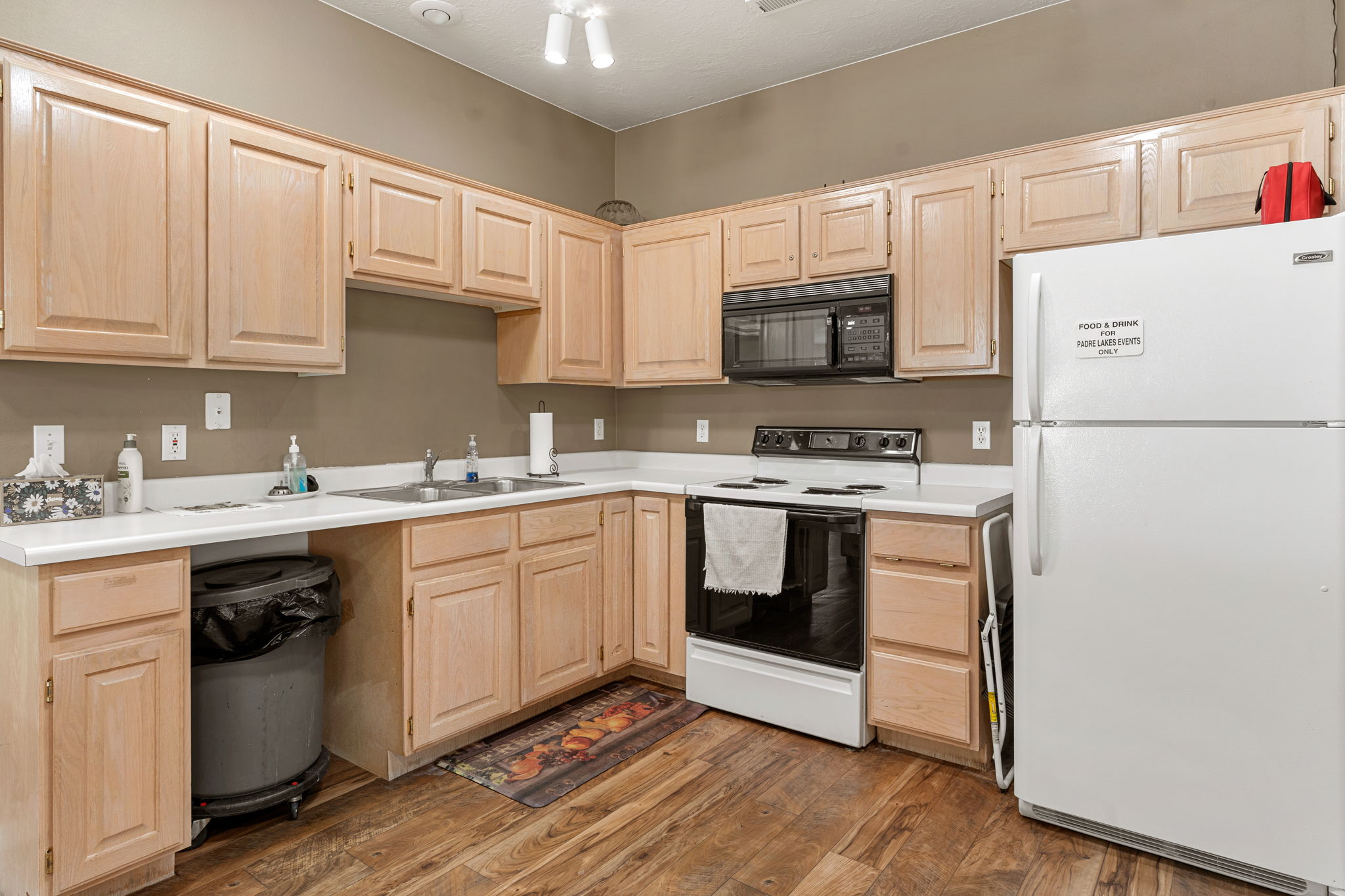 Kitchen featuring light wood cabinetry, white countertops, stainless steel double basin sink, black built-in microwave, and wood-finish flooring