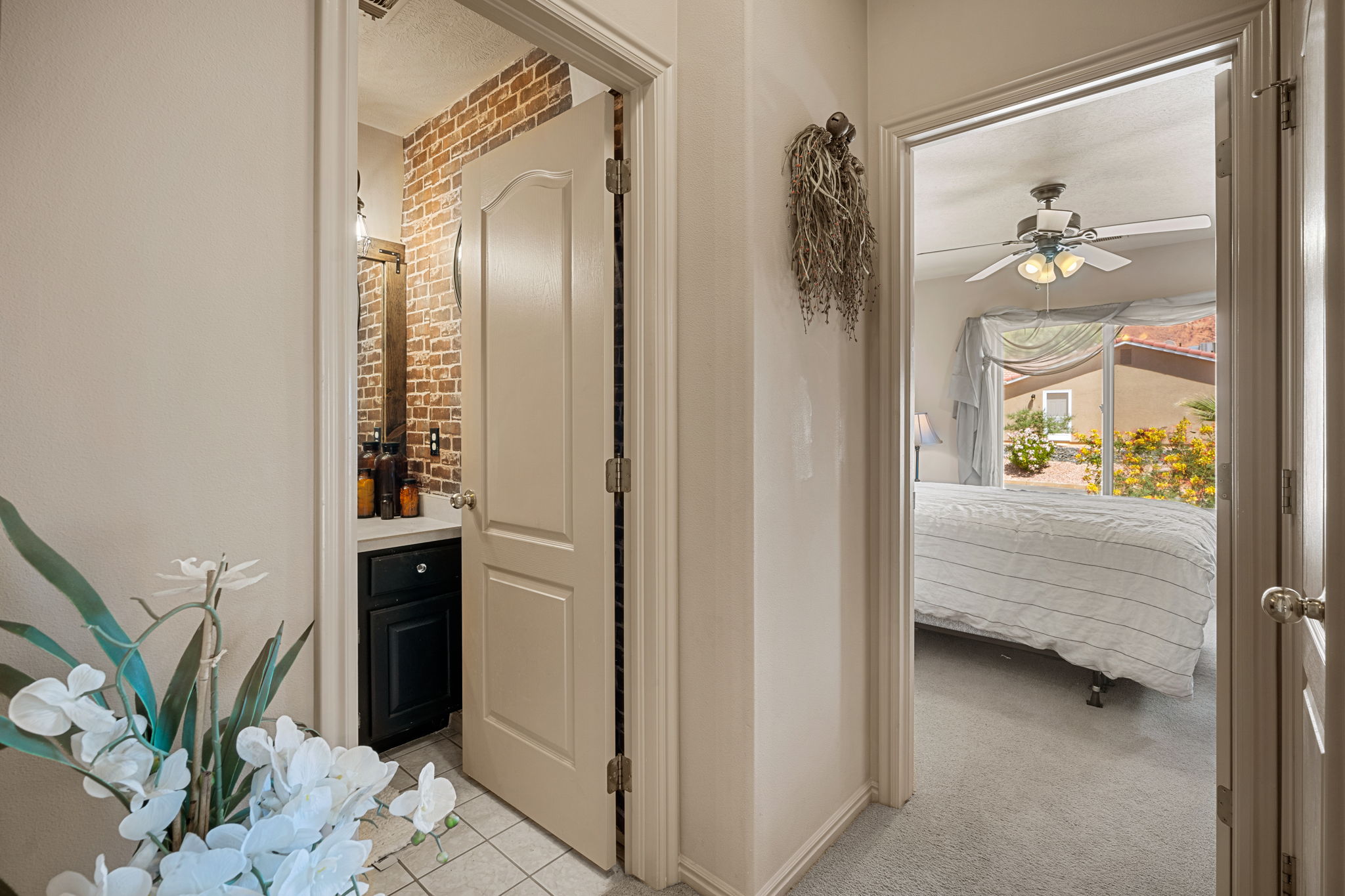 Hallway featuring light-colored walls and doors, with a partial view into a bathroom with a brick-finish accent wall and a dark vanity with a light-colored countertop
