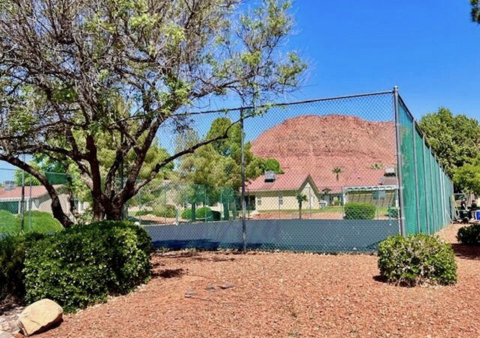 Tennis court with green chain-link fencing, surrounded by mature landscaping and a natural rock backdrop