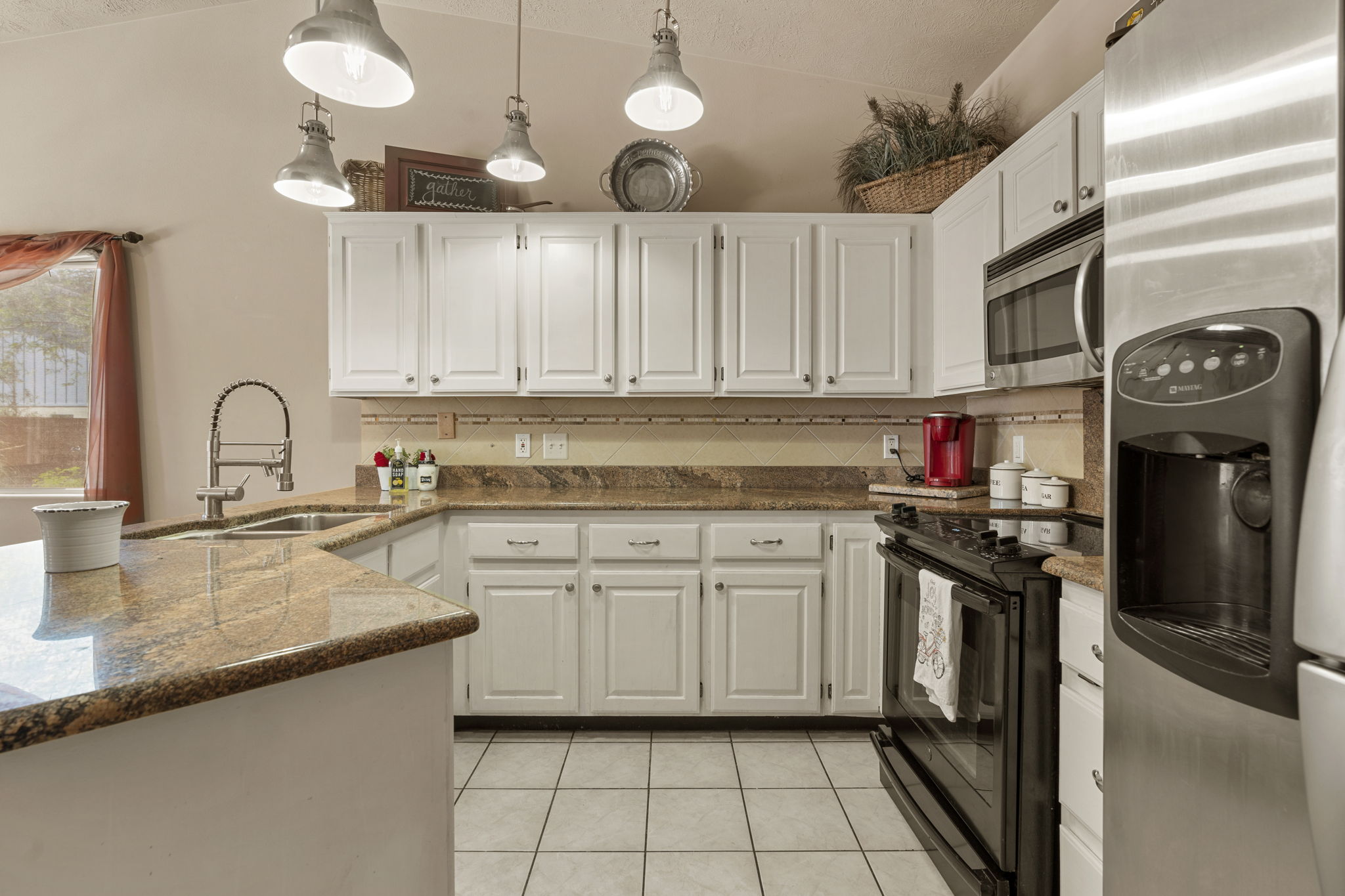 Kitchen featuring white cabinetry, light-toned countertops, a stainless steel sink with a high-arc faucet, white tile flooring, and integrated stainless steel appliances