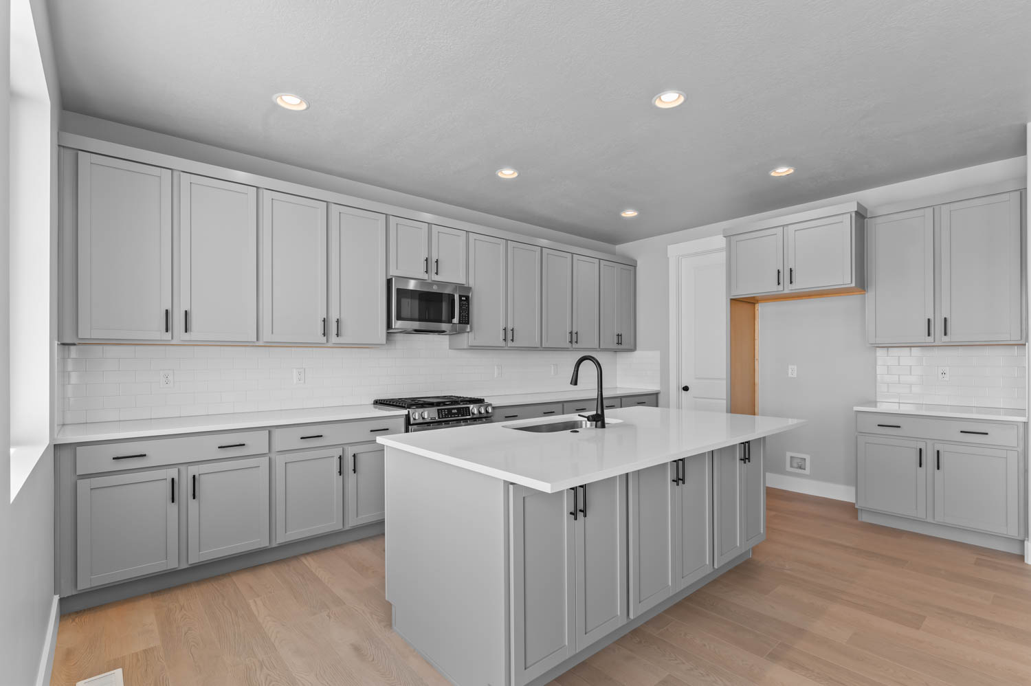 Kitchen featuring a central island with an integrated sink, light gray shaker-style cabinetry with matte black hardware, white subway tile backsplash, stainless steel appliances, and light wood-finish flooring