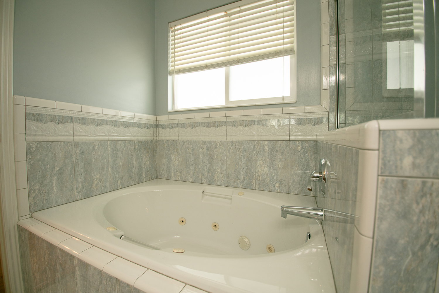 Corner jetted tub with tiled surround, light blue painted walls, and window with blinds