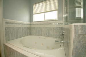 Corner jetted tub with tiled surround, light blue painted walls, and window with blinds