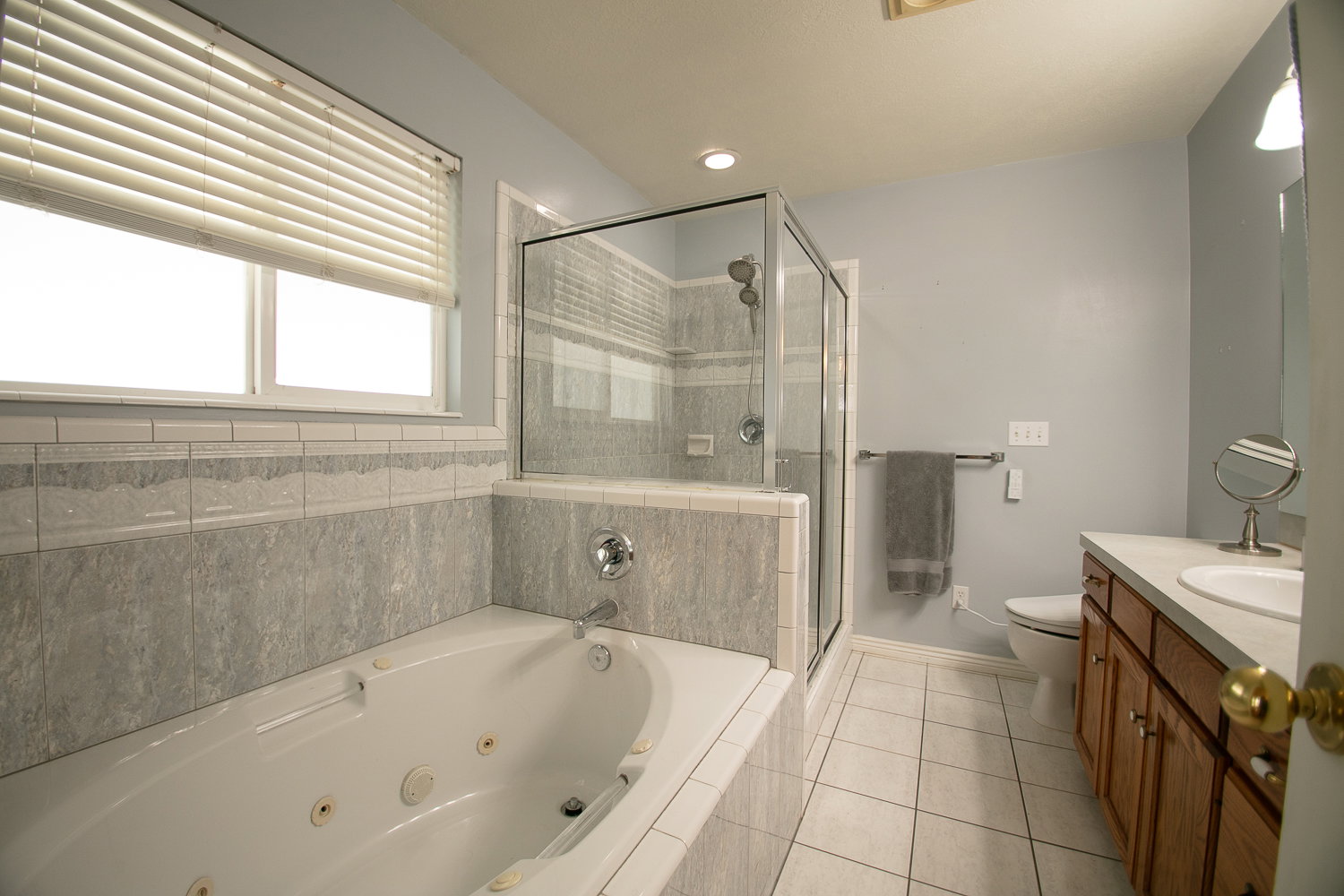 Primary bathroom with Jetted tub with tiled surround, glass-enclosed shower, white tile flooring, double-pane window with blinds, and a wood-finish vanity with countertop sink