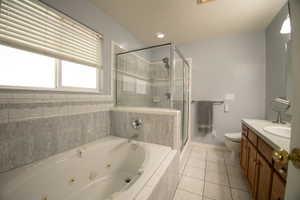 Primary bathroom with Jetted tub with tiled surround, glass-enclosed shower, white tile flooring, double-pane window with blinds, and a wood-finish vanity with countertop sink