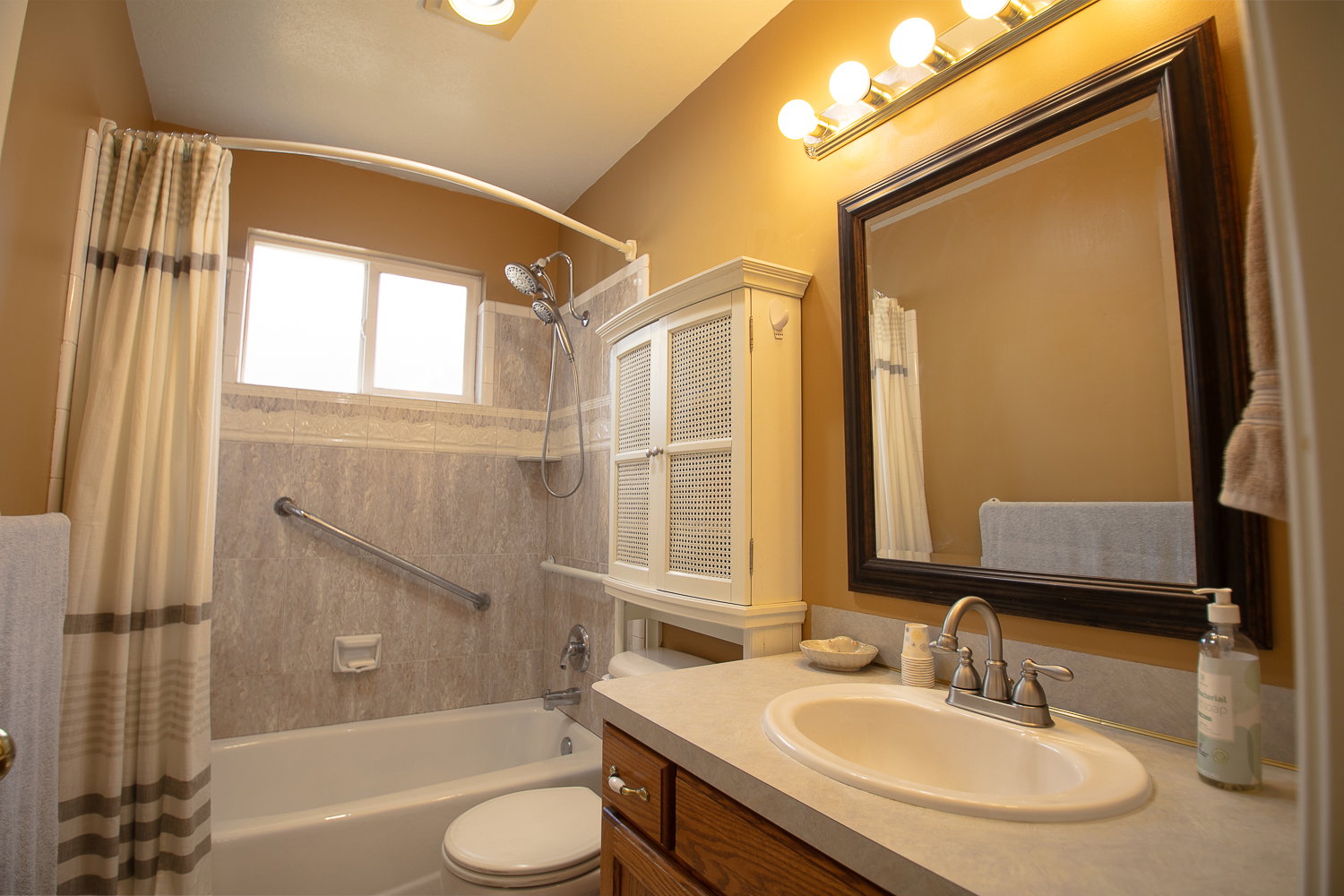 Bathroom featuring a combination shower and tub with neutral-toned wall surround, an overhead window, a wood-finish vanity with a built-in sink, and a framed mirror with overhead lighting