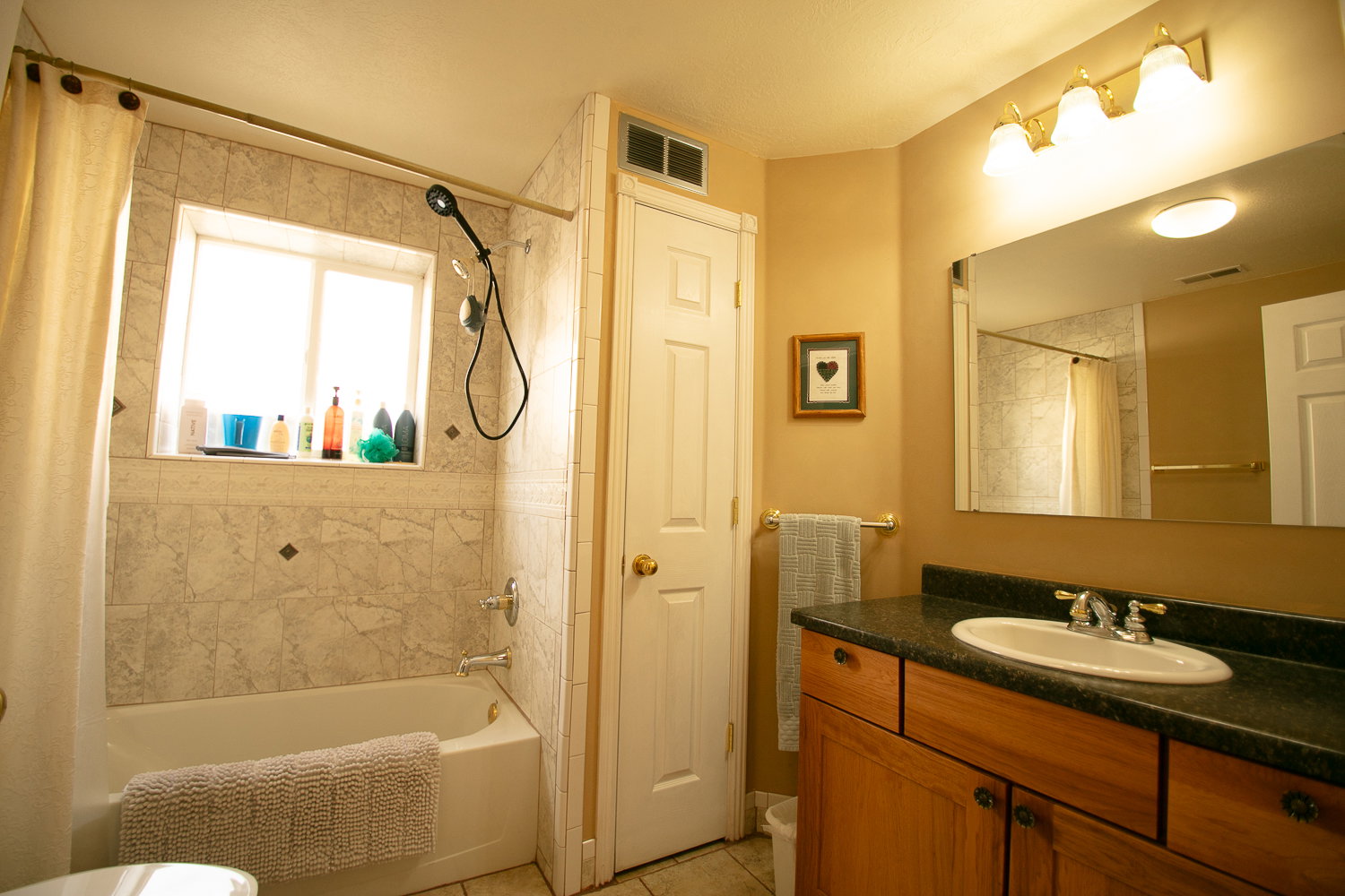Bathroom featuring a tiled tub and shower surround with an inset window, a wood-finish vanity with a dark countertop, a white oval sink, and a rectangular mirror with a three-light fixture