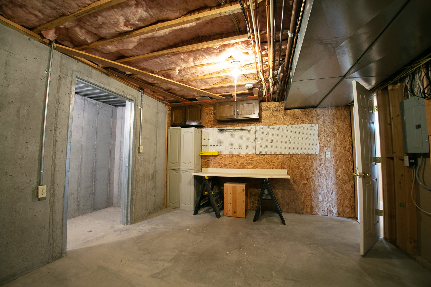 Unfinished basement area featuring concrete walls, exposed ceiling joists with insulation, and a utility workbench with shelving
