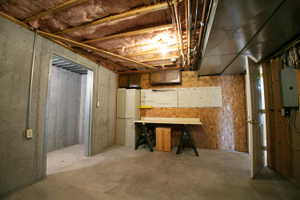 Unfinished basement area featuring concrete walls, exposed ceiling joists with insulation, and a utility workbench with shelving