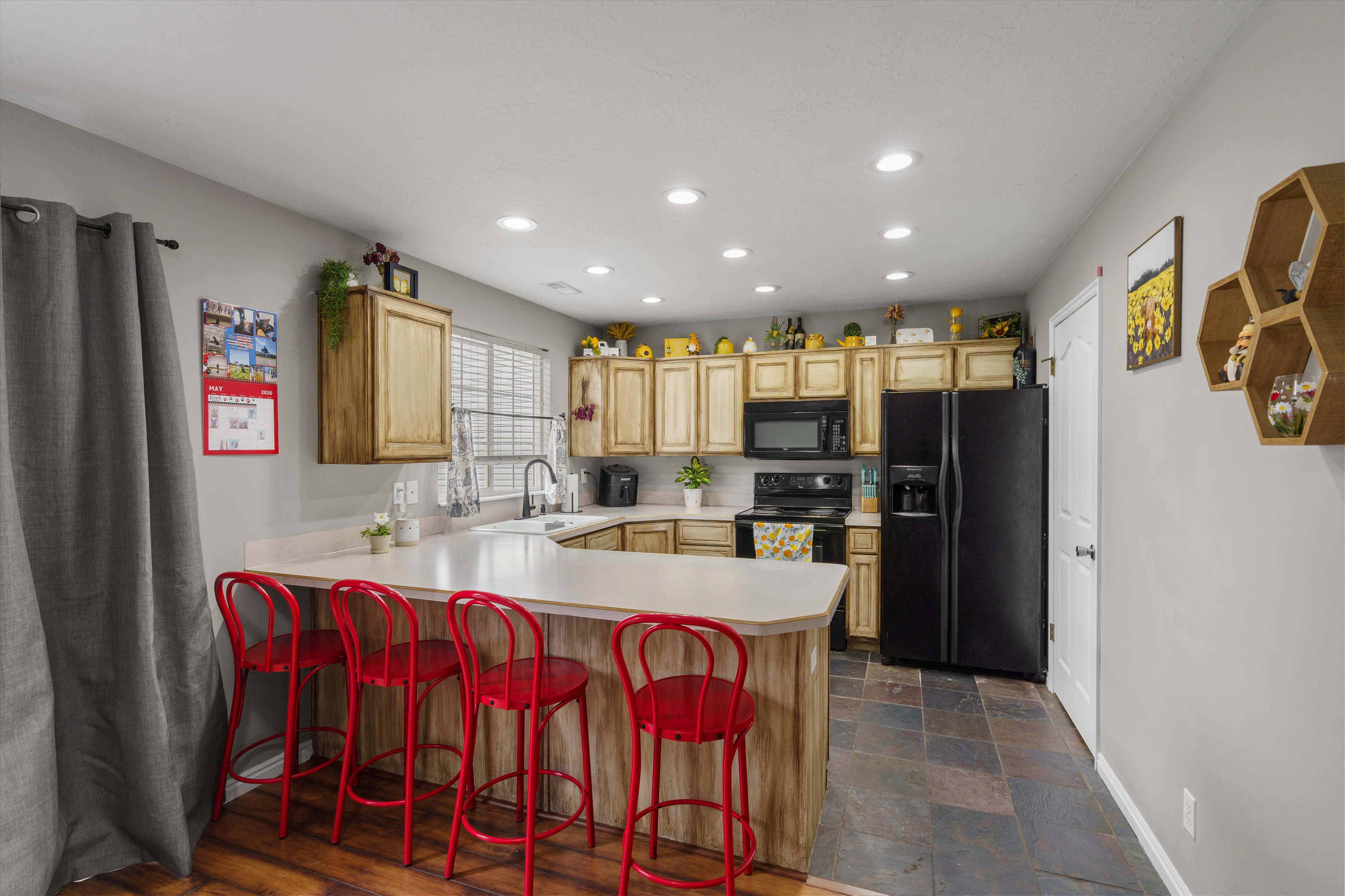 Kitchen featuring a U-shaped island with a light-toned countertop, wood-finish cabinetry, black appliances, recessed lighting, and slate-style floor tiling. Oversized pantry