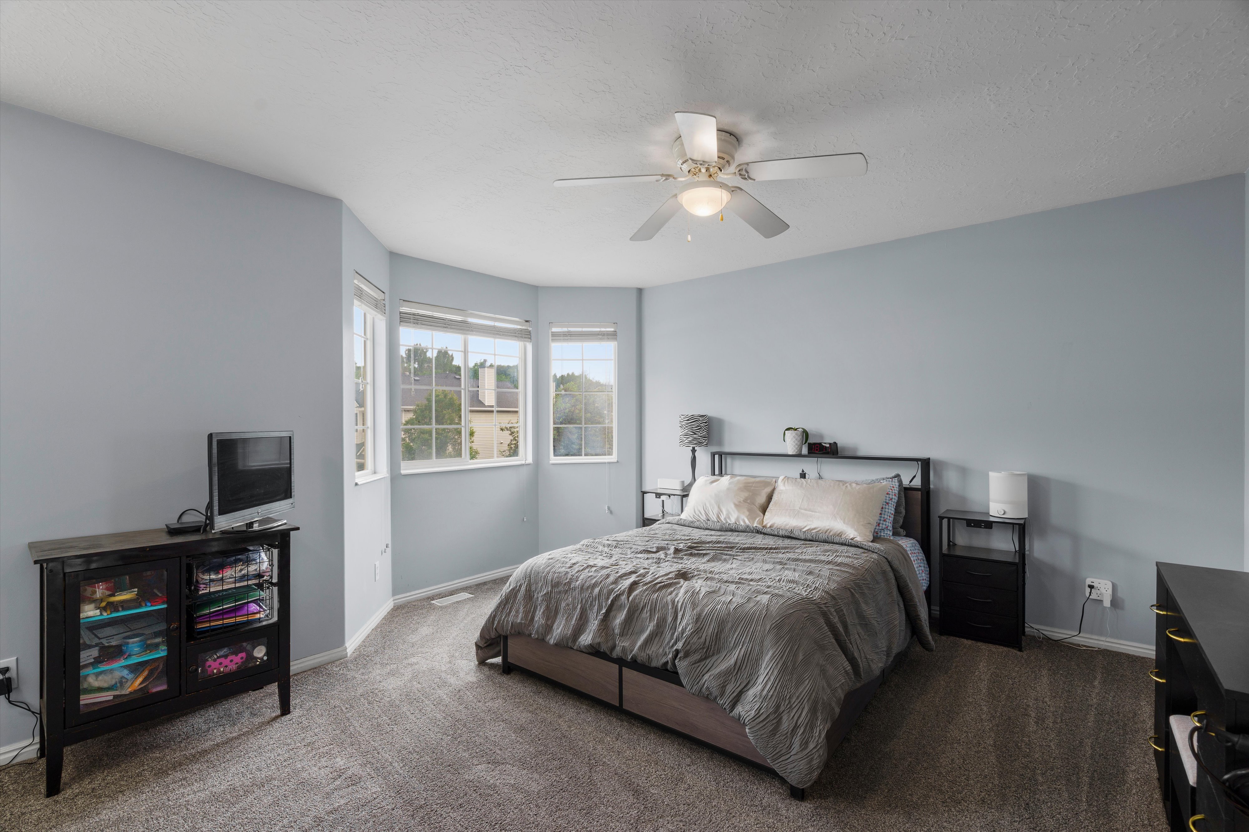 Oversized primary bedroom featuring a bay window with multiple panes, fan with integrated lighting, and white trim