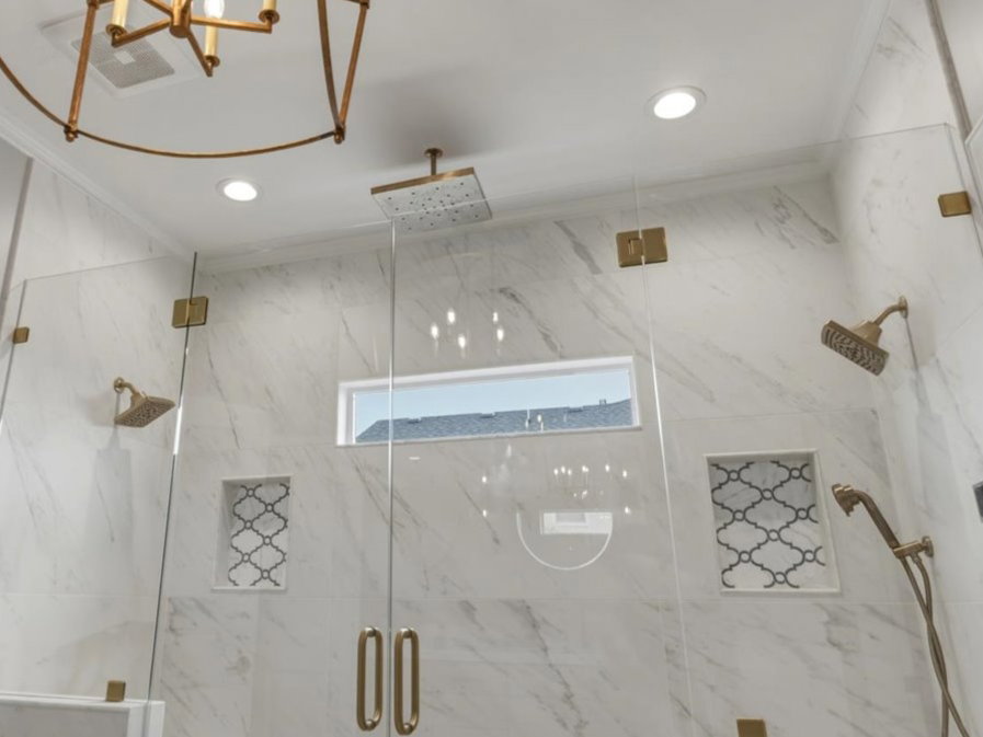 Spacious shower enclosure featuring full-height marble-look tile, frameless glass panels with brass-finish hardware, and a built-in bench
