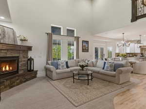 Spacious living area featuring a stacked stone fireplace with a wood mantel, high ceilings, large windows with white louvered shutters, and a partial view of a kitchen with light wood flooring