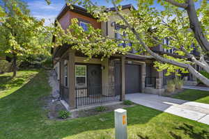 Contemporary multi-level residence featuring a covered porch with metal railing, an integrated garage, and a sloped grassy yard with mature trees