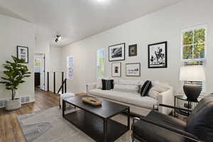 Bright living area featuring wood-finish flooring, white walls, multiple windows with blinds, track lighting, and a modern stair railing