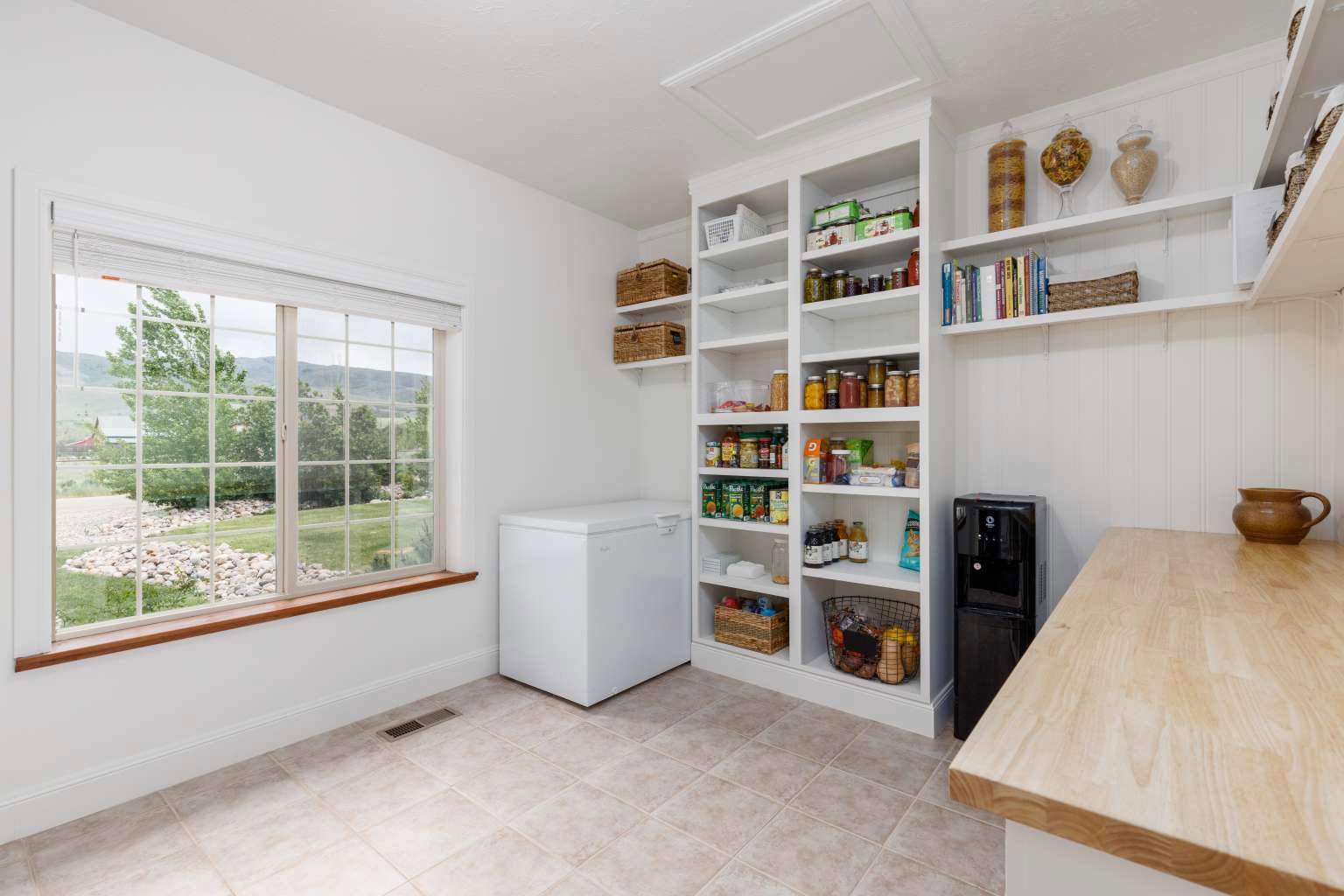 Spacious pantry featuring extensive built-in shelving, a large window with natural light, tiled flooring, and a wood-finish countertop
