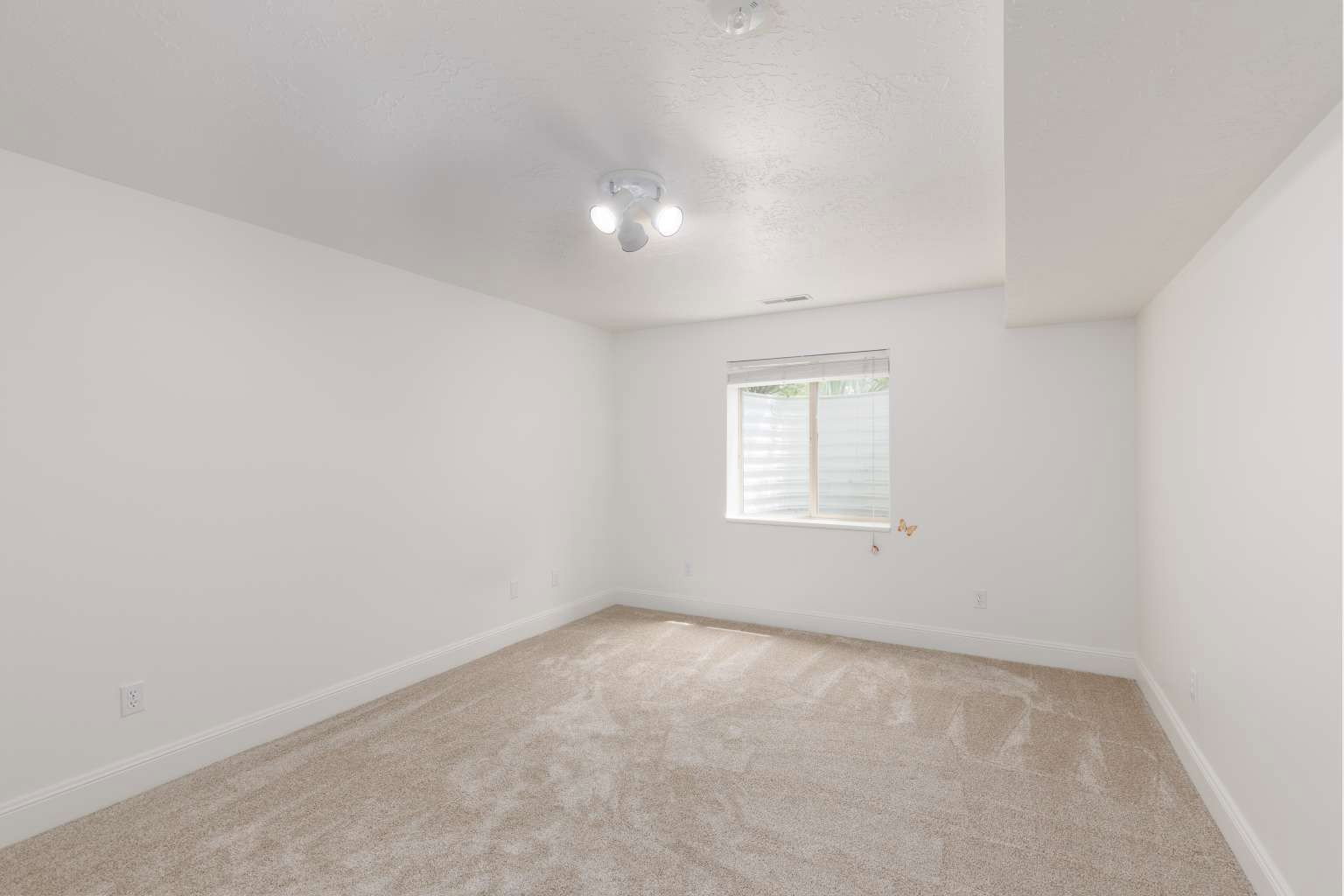 Neutral-toned carpeting extends throughout this room featuring a window with horizontal blinds and an overhead three-light fixture