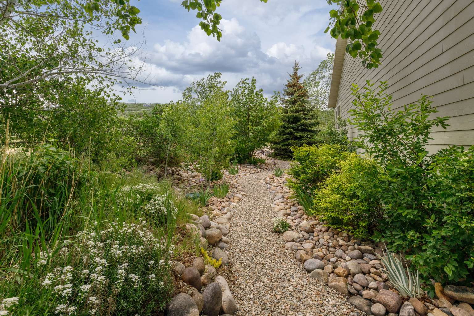 Gravel pathway bordered by river rocks, leading through dense foliage and flowering plants