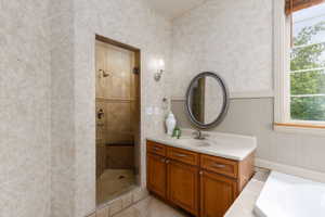 Bathroom featuring a glass-enclosed shower with tiled walls and built-in bench, a wooden vanity with a light-colored countertop and integrated sink, a framed oval mirror, a window with natural light, and tiled flooring