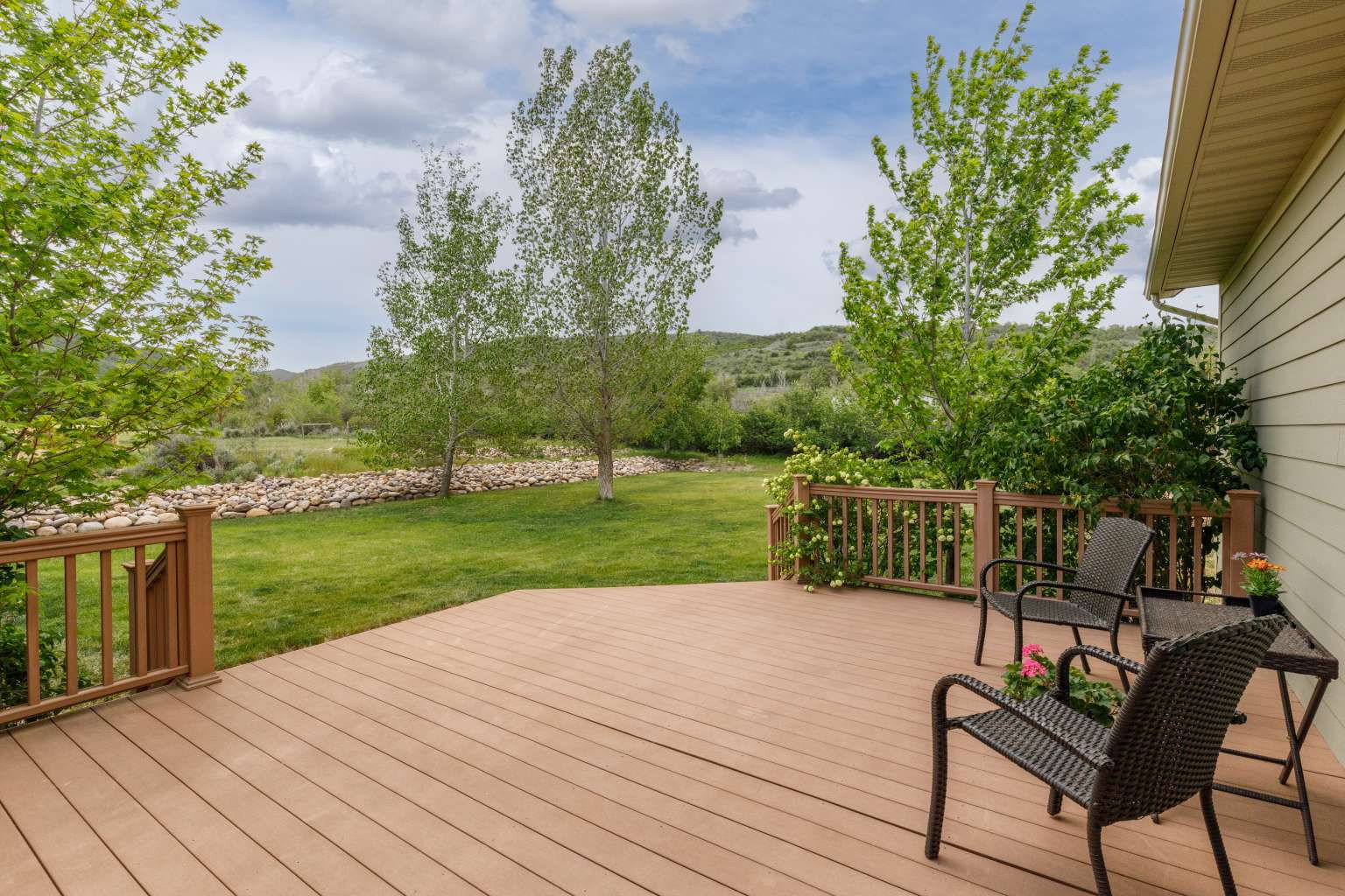 Spacious outdoor deck with composite decking and wood railings, overlooking a manicured lawn, mature trees, and natural rock features