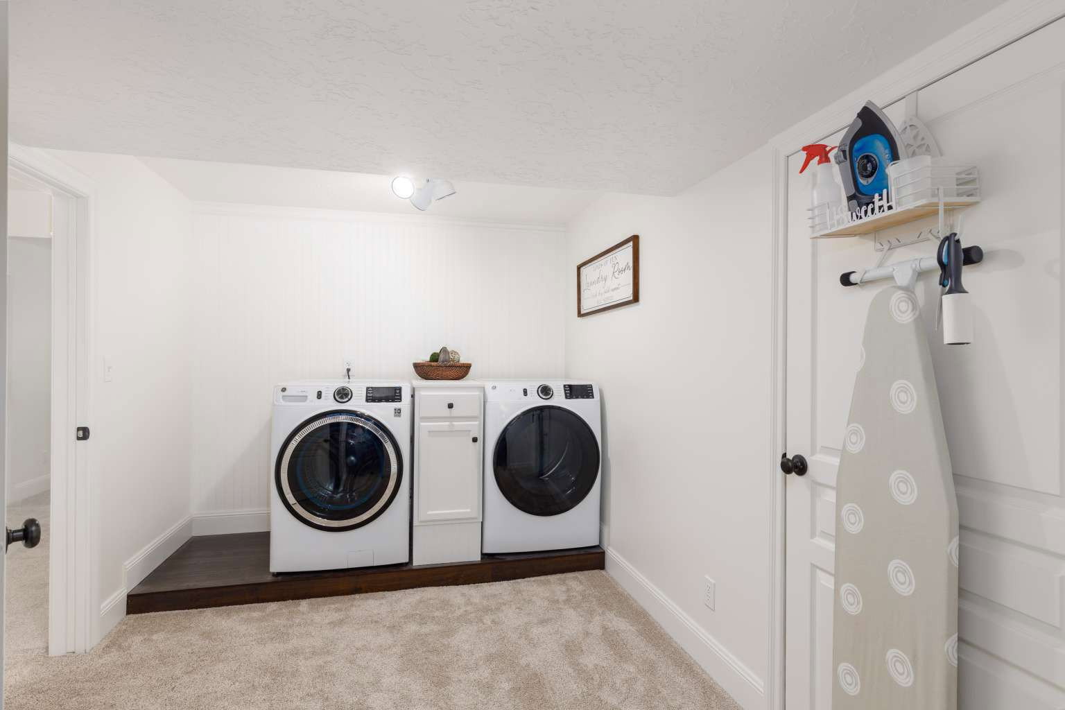 Dedicated laundry area featuring a raised wood-finish platform for appliances, white paneled walls, and soft neutral carpeting