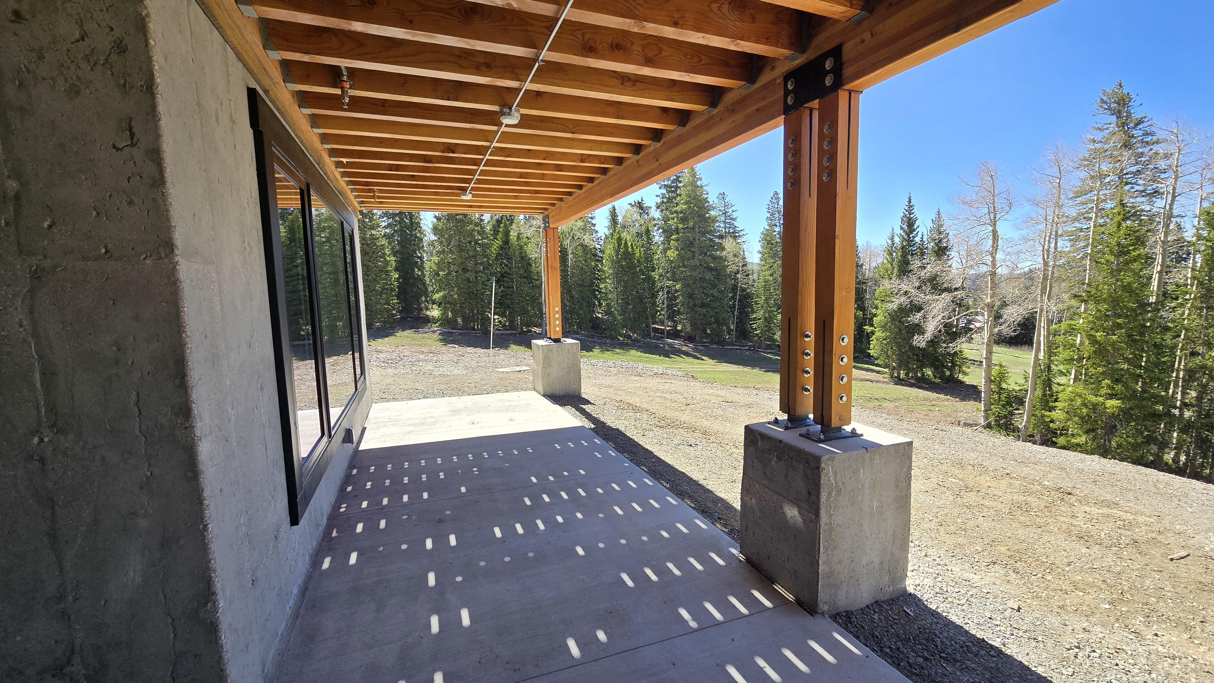 Covered concrete patio featuring exposed wood beams, concrete support columns, and large black-framed windows
